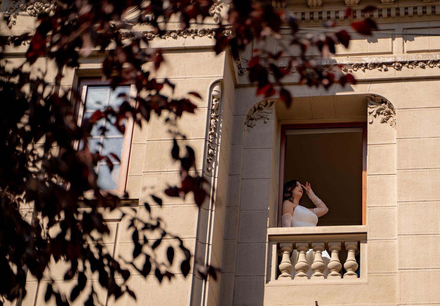 Woman standing on ornate building balcony looking away, framed by red autumn leaves in foreground.