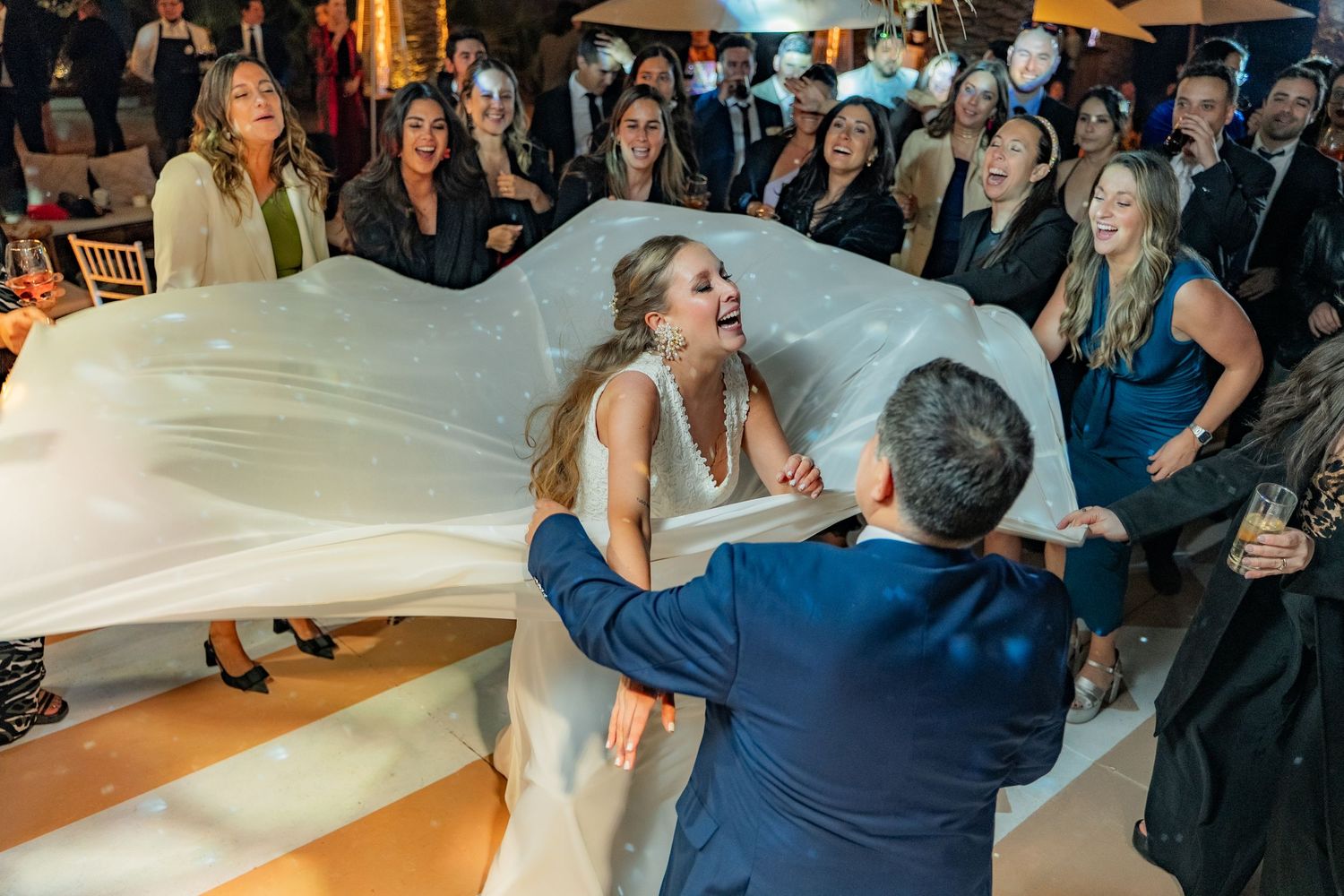 Wedding couple dances while guests hold up flowing white dress on dance floor.