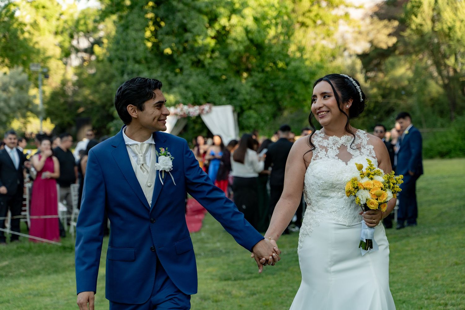 A joyful wedding couple walks hand in hand on a lawn with guests in the background.