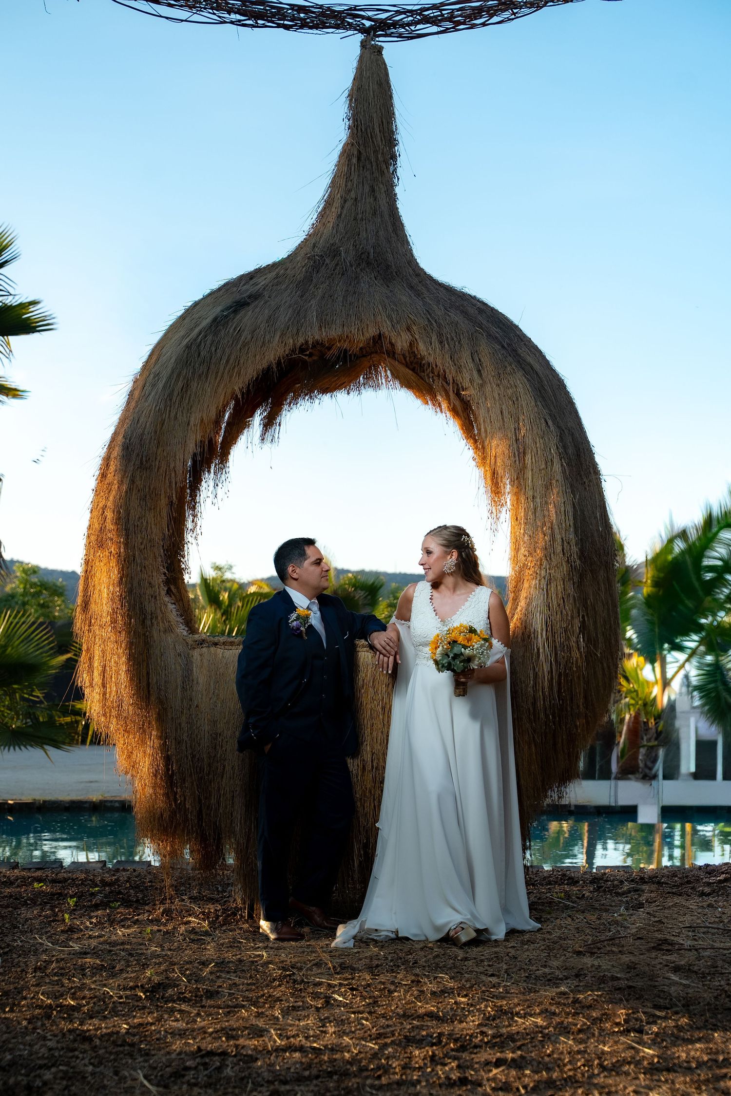 Wedding couple stands beneath unique thatched arch structure at outdoor venue.