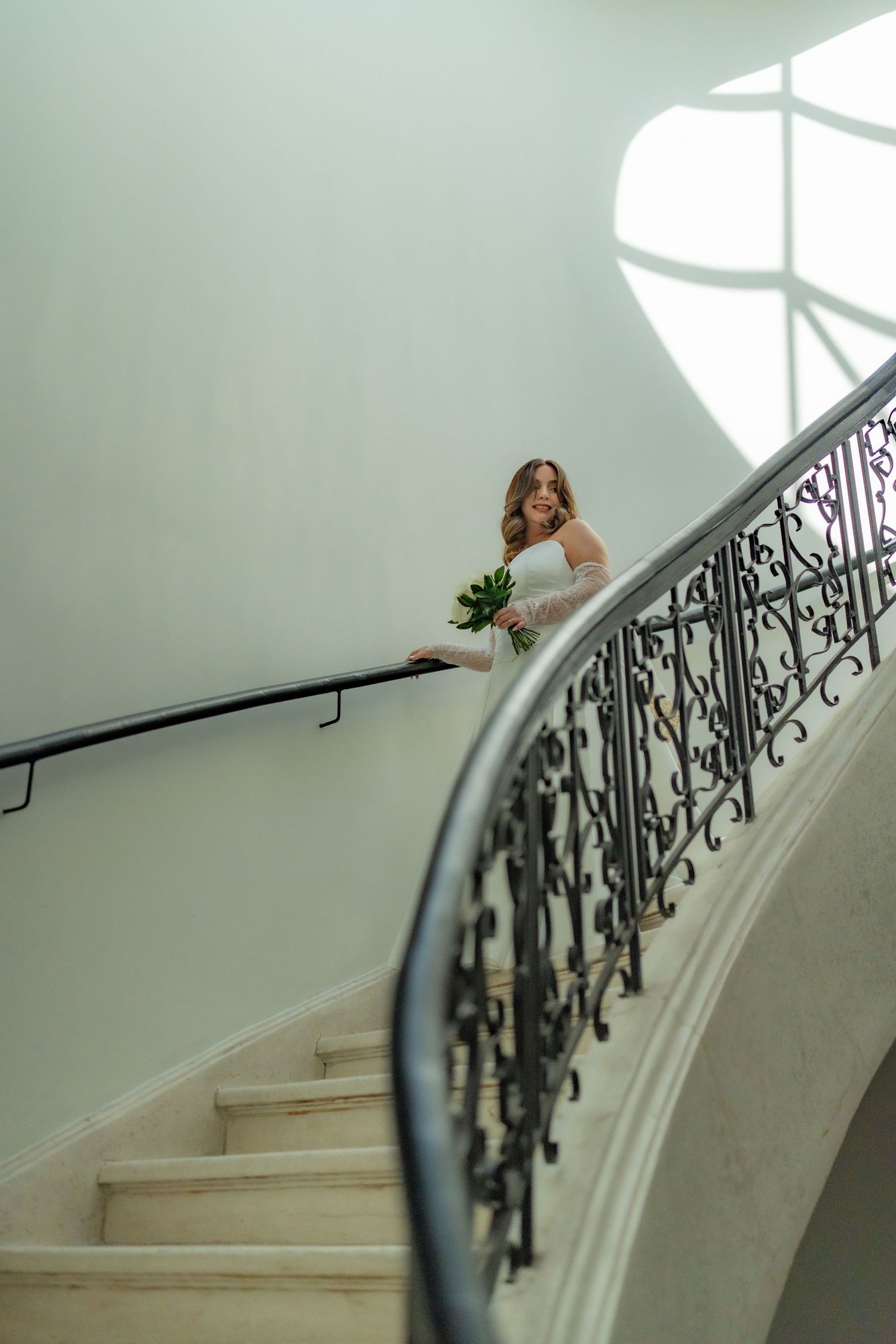 Bride in white wedding dress holding bouquet posing on grand curved staircase with iron railing.
