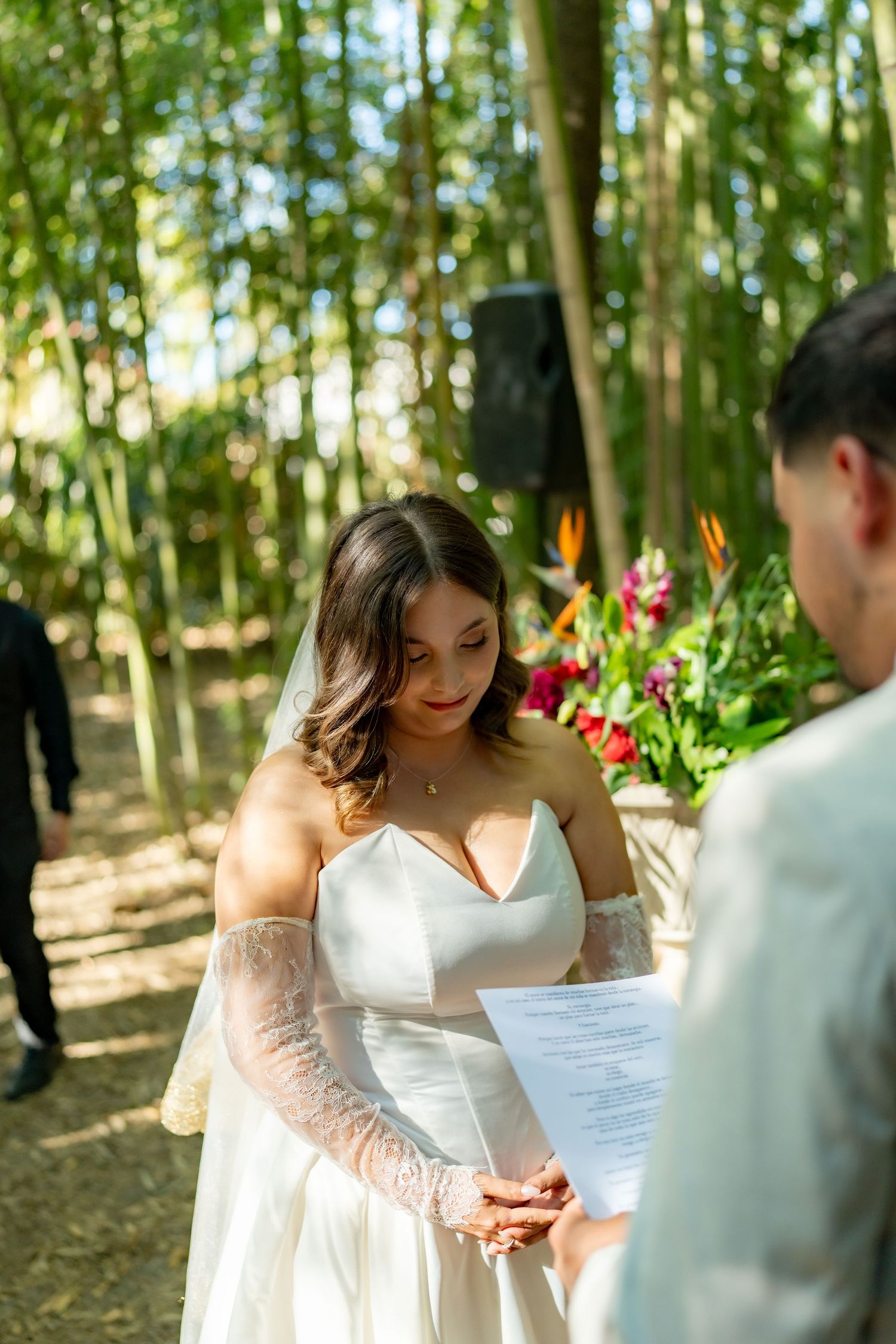 Bride in strapless white gown with lace sleeves reading wedding vows during outdoor bamboo ceremony.