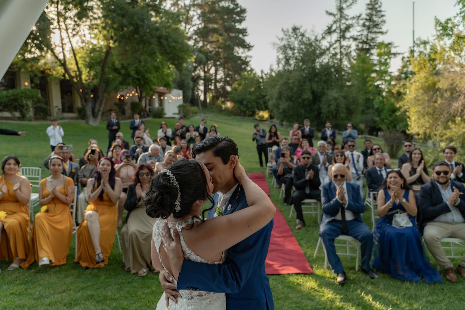 A group of wedding guests seated outdoors in blue attire enjoy a summer evening celebration in a garden setting.