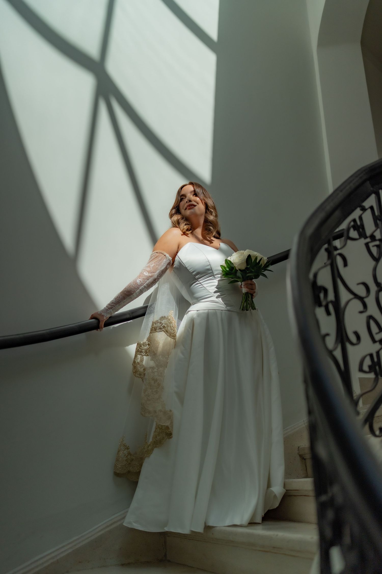Bride in white gown with lace sleeves holds bouquet while posing on a grand staircase with decorative iron railing.
