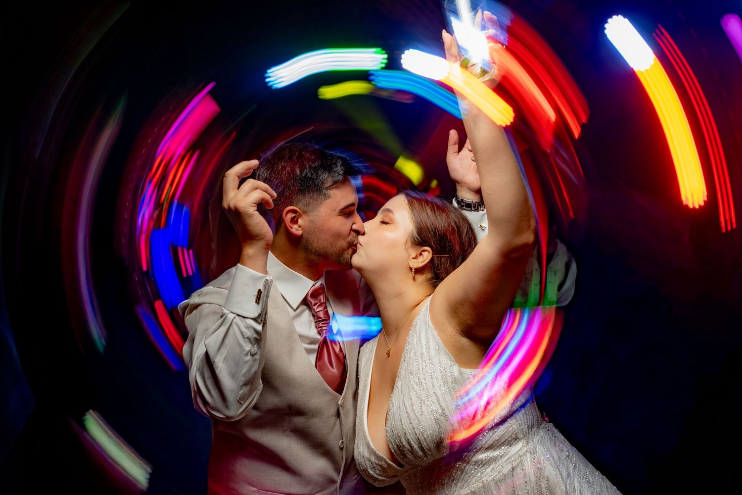 Wedding couple sharing a romantic kiss surrounded by colorful swirling neon lights in a dark reception venue.