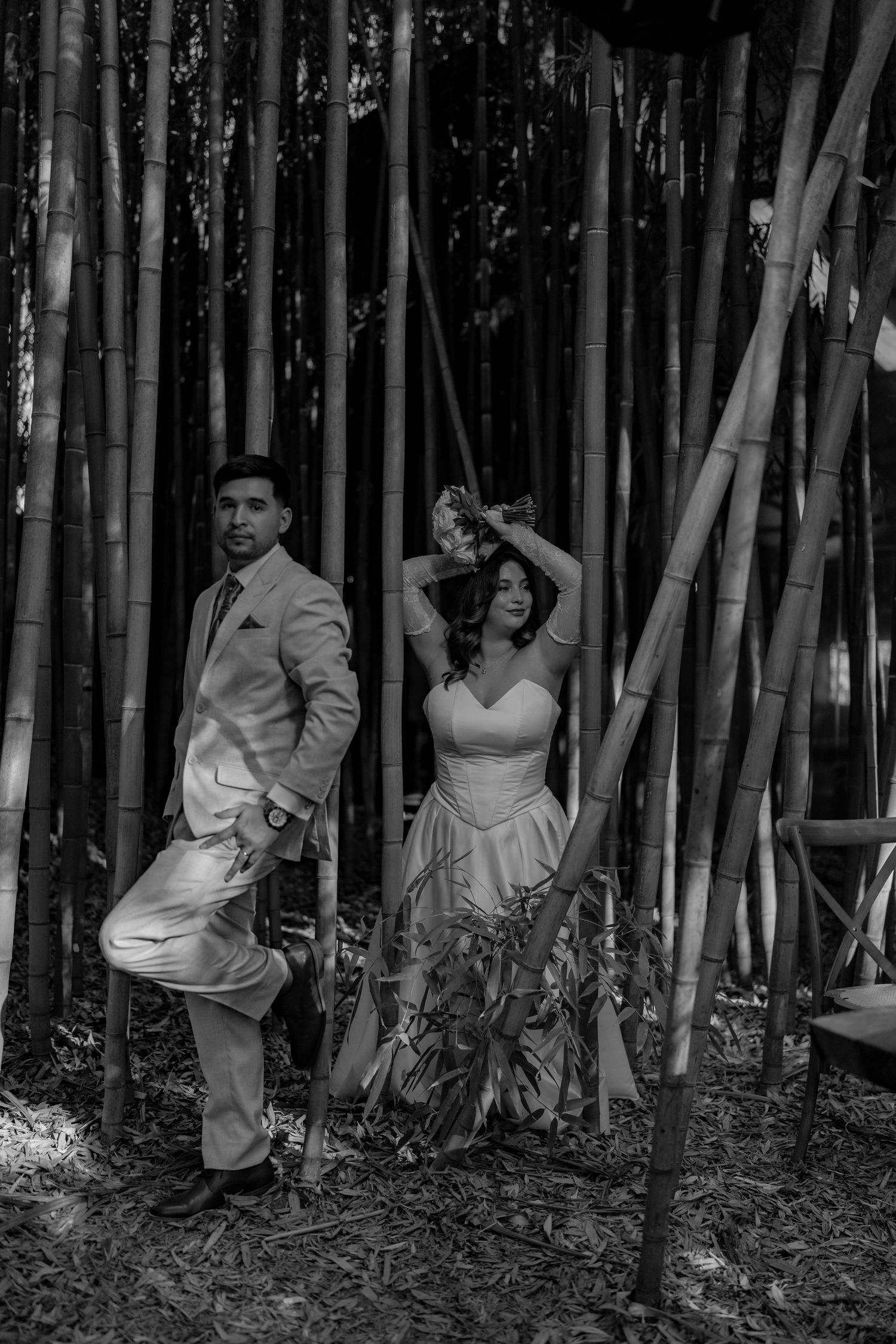 Black and white photo of bride and groom posing playfully among tall bamboo trees.