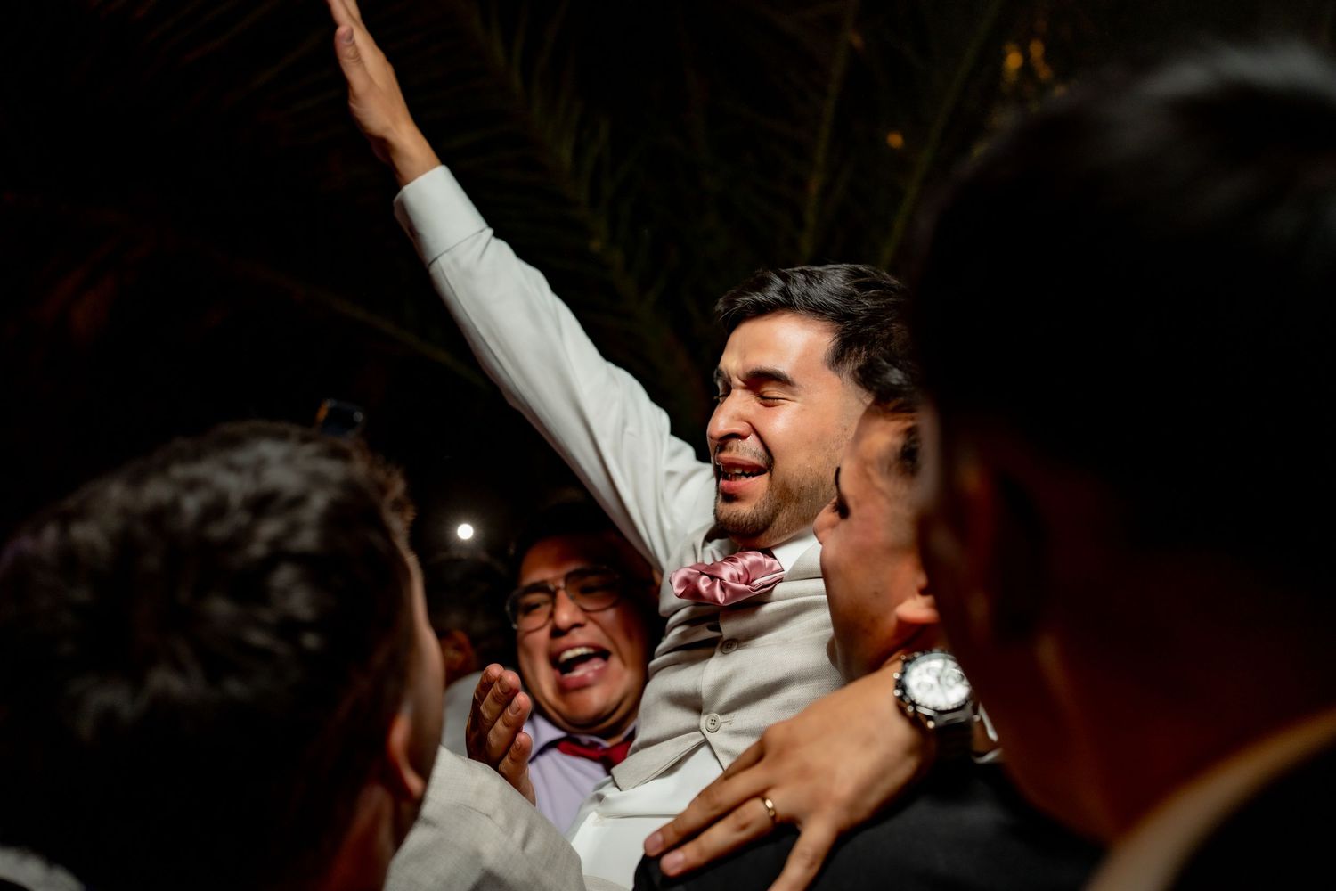 Groom lifted by cheering guests with arms raised in celebration at lively outdoor wedding reception.