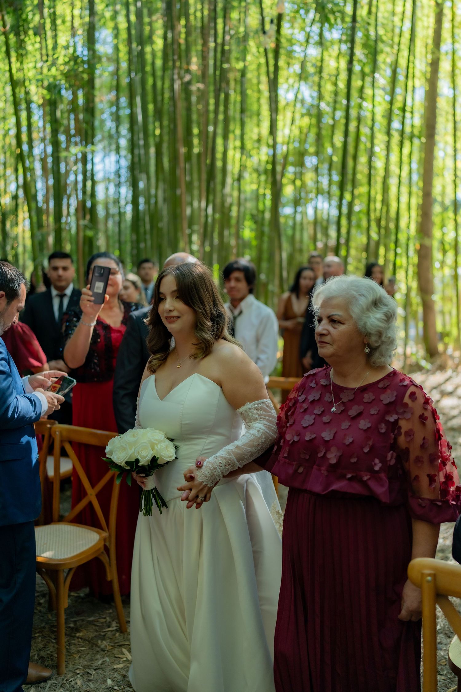 Bride in white gown walks down bamboo forest aisle escorted by older woman in burgundy dress.
