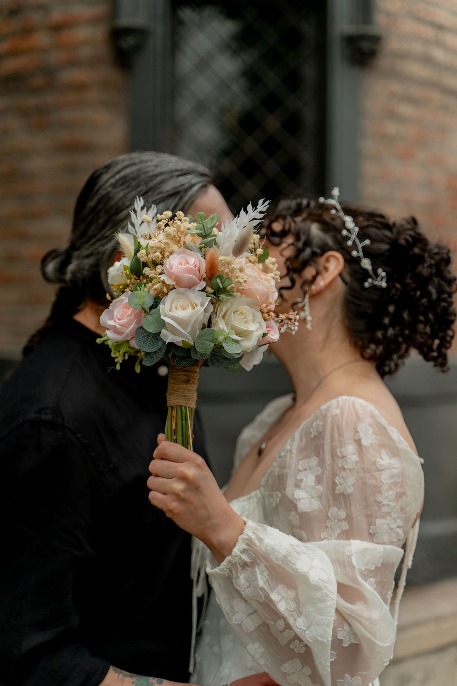 Bride in delicate lace wedding dress holds a romantic bouquet of pink and white roses against a brick wall backdrop.