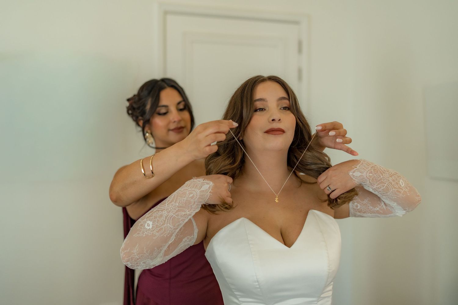Bridesmaid in burgundy dress helping bride in white gown put on a delicate necklace before wedding ceremony.