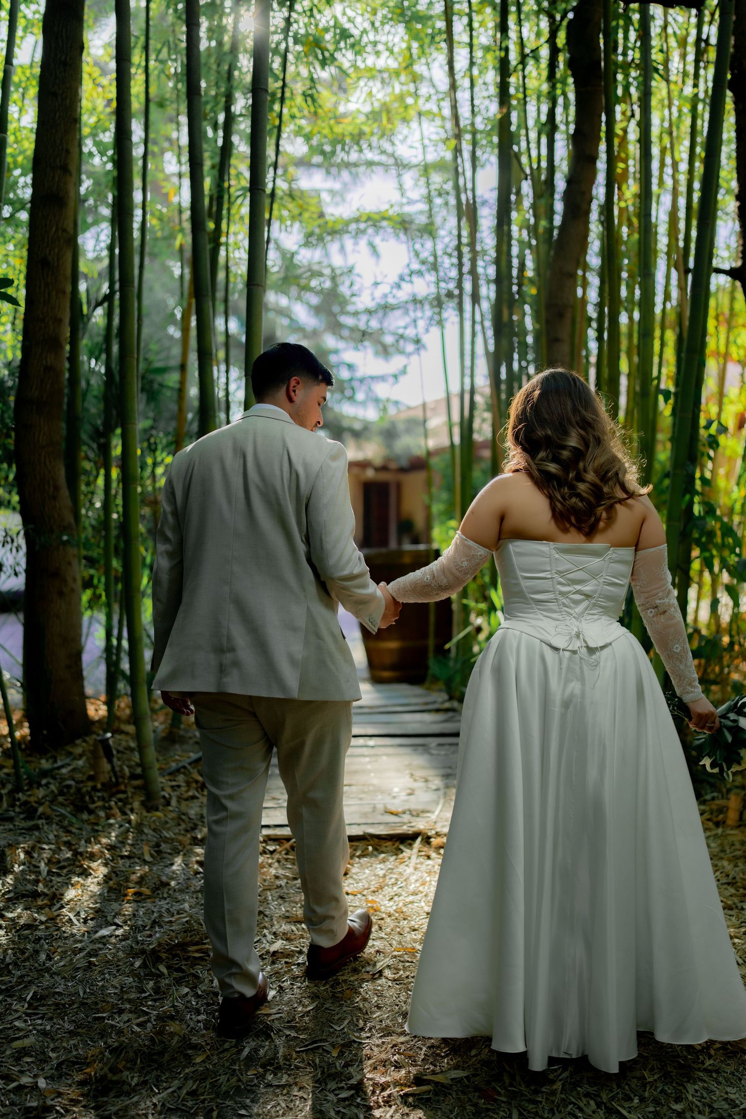 Newlywed couple holding hands walking through lush bamboo forest path after wedding ceremony.