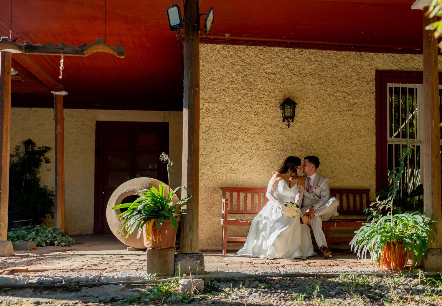 Bride and groom share a romantic kiss on a rustic porch bench surrounded by lush greenery.