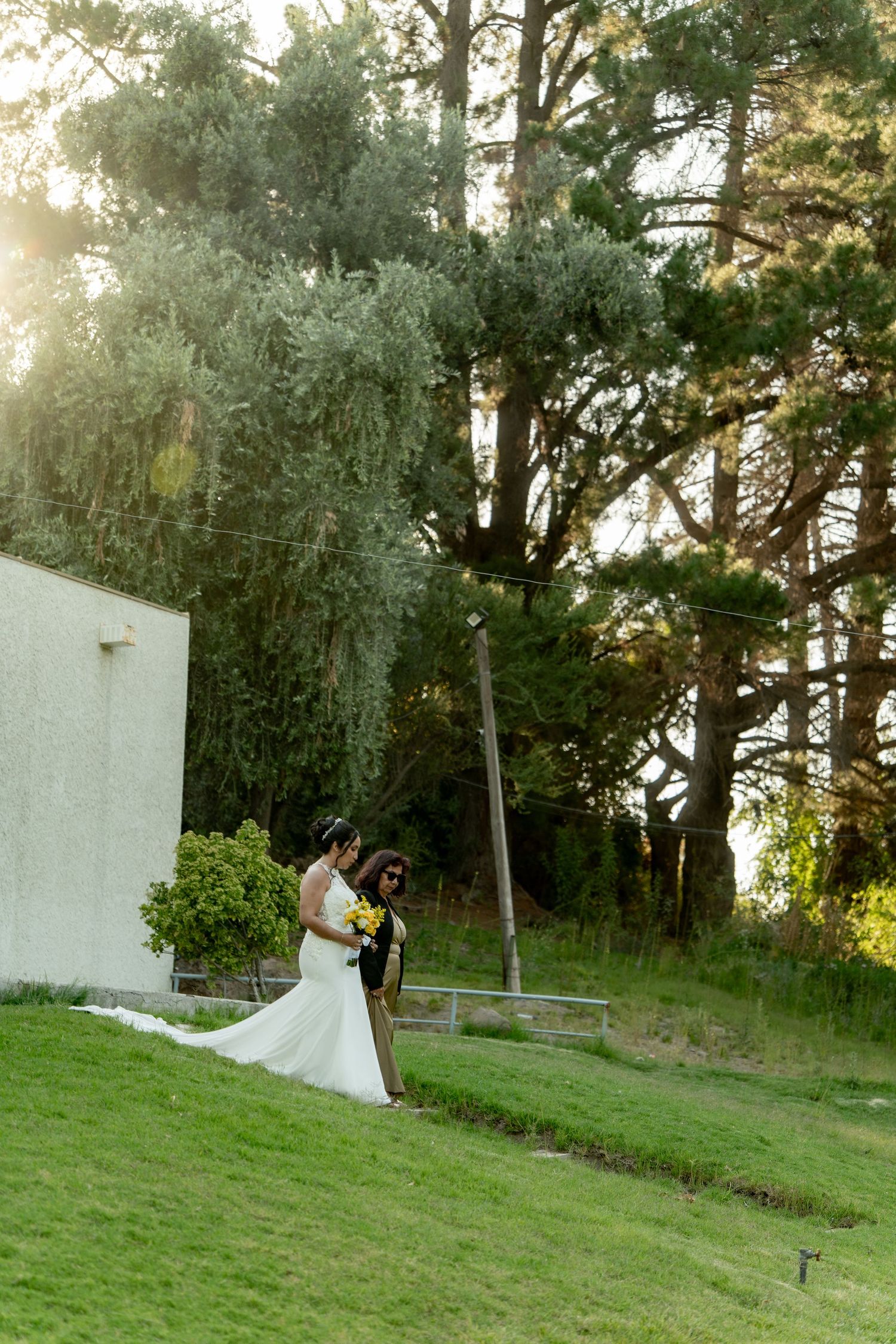 Wedding couple posing for photos on grassy hill with white wall backdrop at sunset.