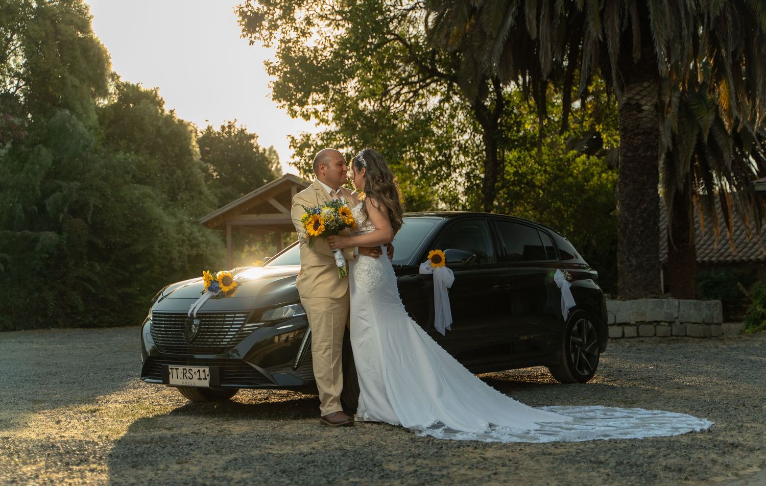A wedding couple poses with a luxury car during golden hour under palm trees.