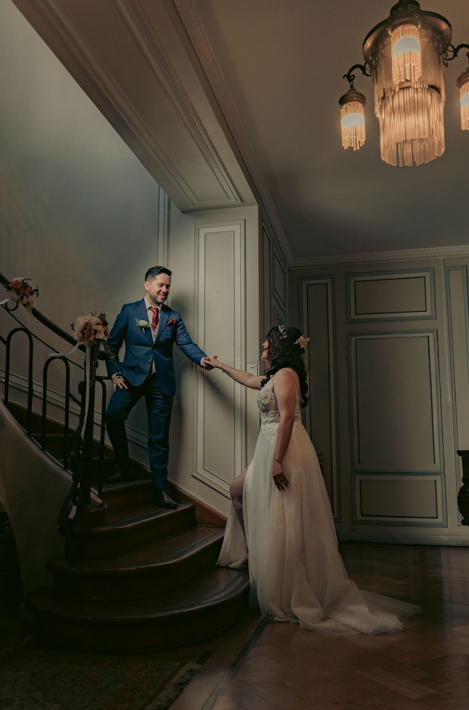 Elegant couple poses on a grand staircase in a vintage-style interior.