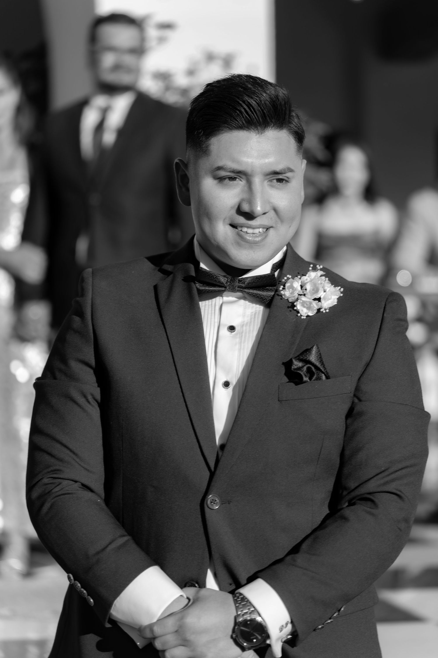 Person in formal black tuxedo smiles during outdoor wedding ceremony.