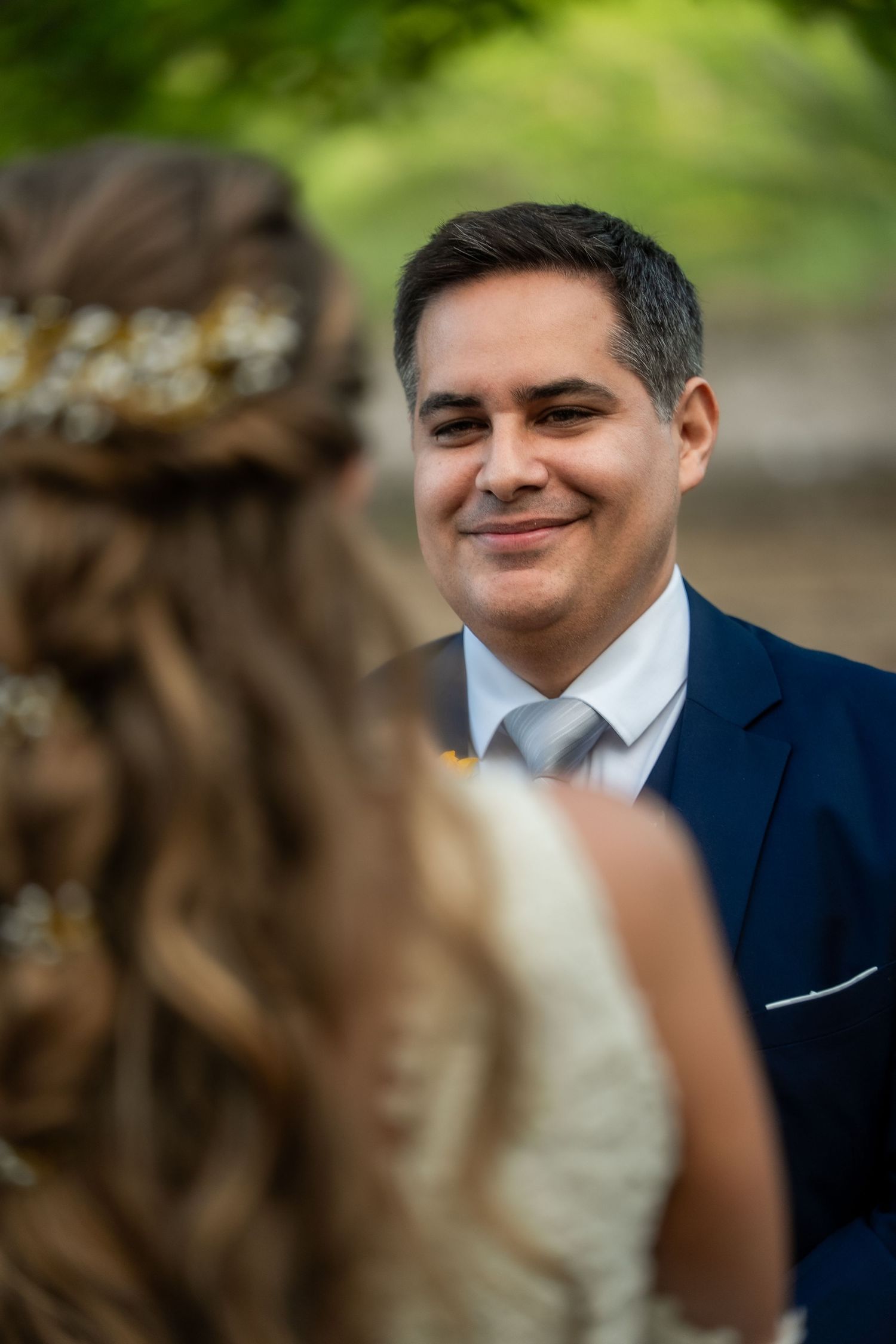 Groom in navy suit smiles warmly during wedding ceremony in garden setting.