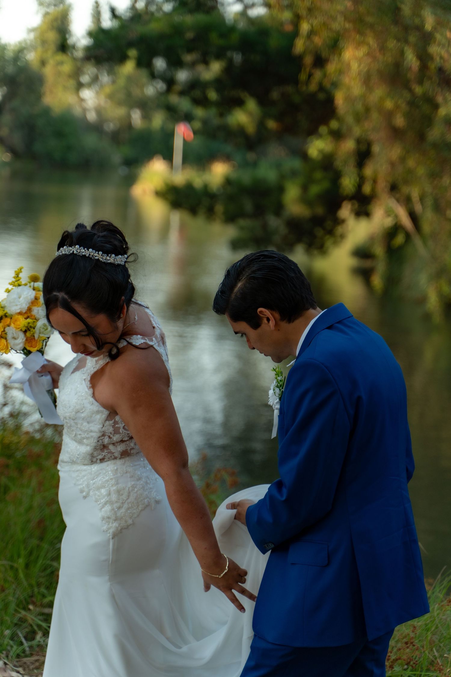 Couple in white dress and blue suit standing by waterside during outdoor celebration.