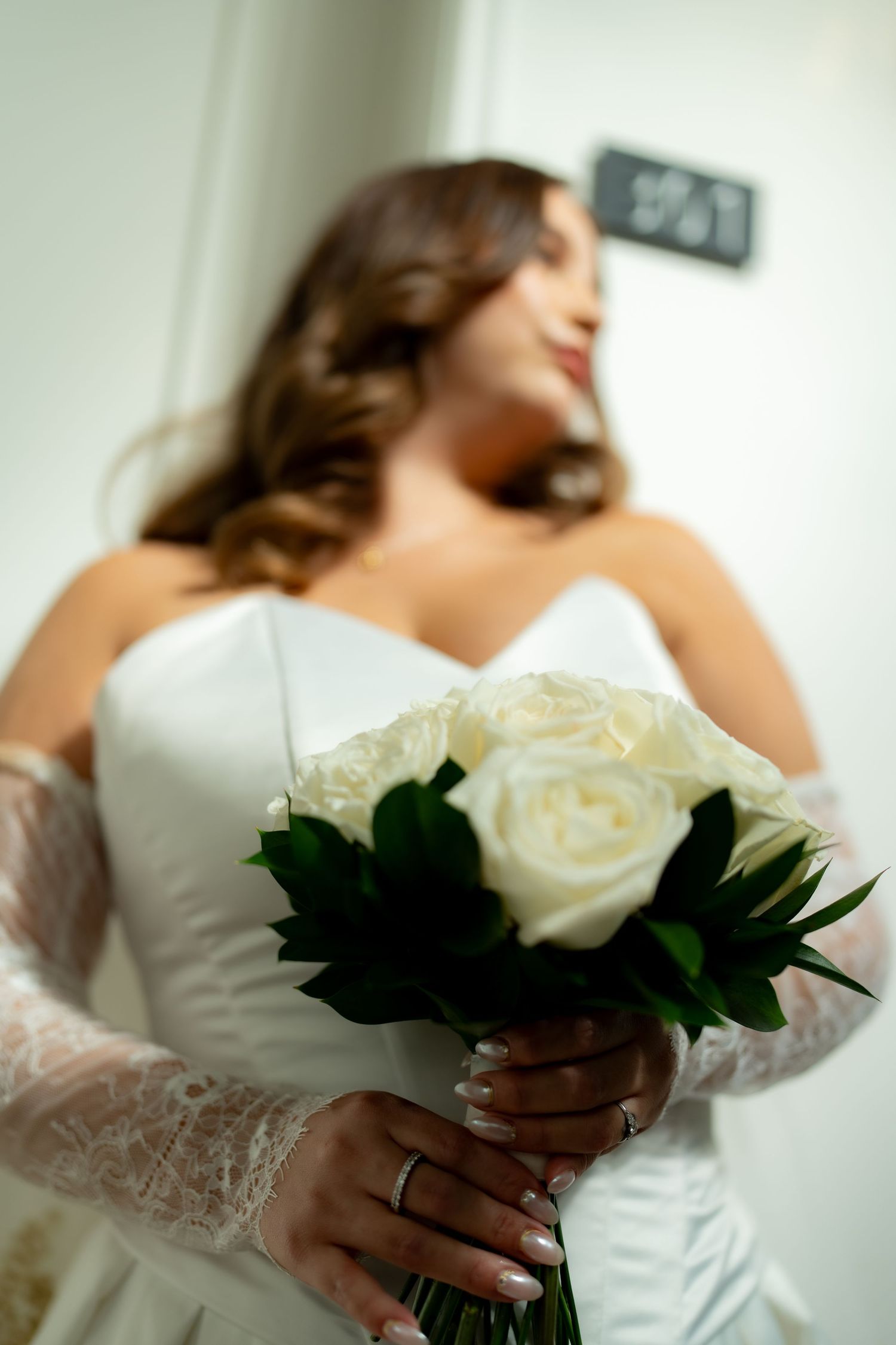 Bride in white strapless gown holds elegant bouquet of white roses with lush green foliage.