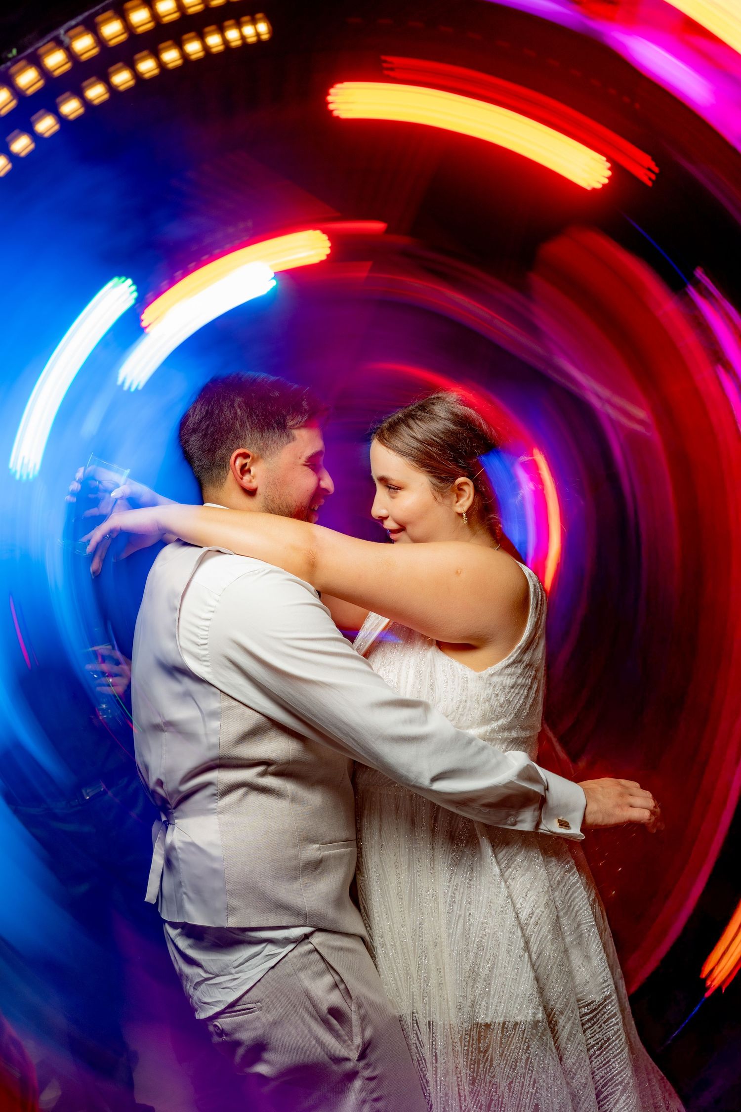Bride and groom sharing first dance embraced in swirling colorful neon lights at wedding reception.