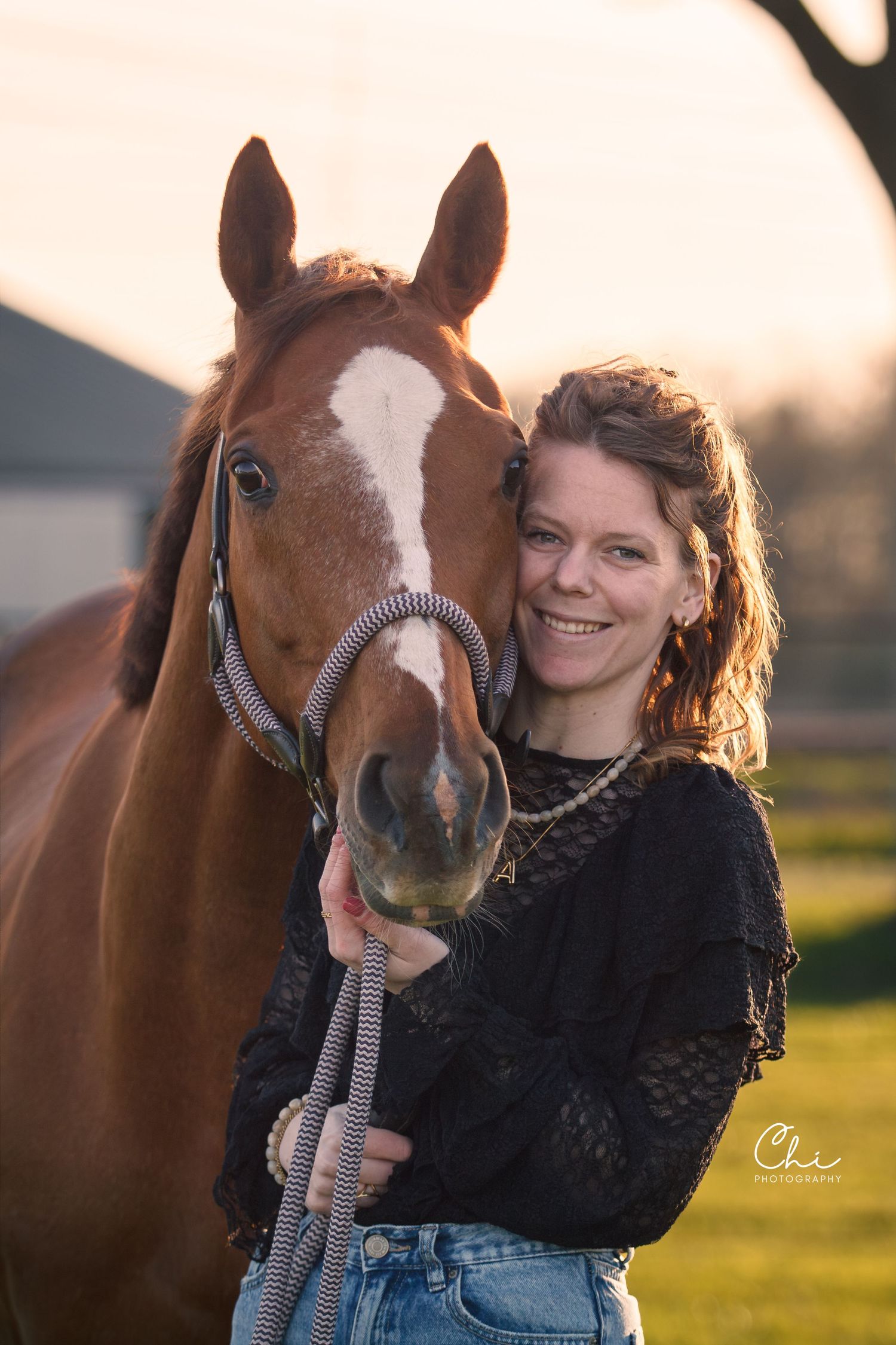 A happy woman poses closely with a chestnut horse wearing a rope halter during a warm golden sunset.