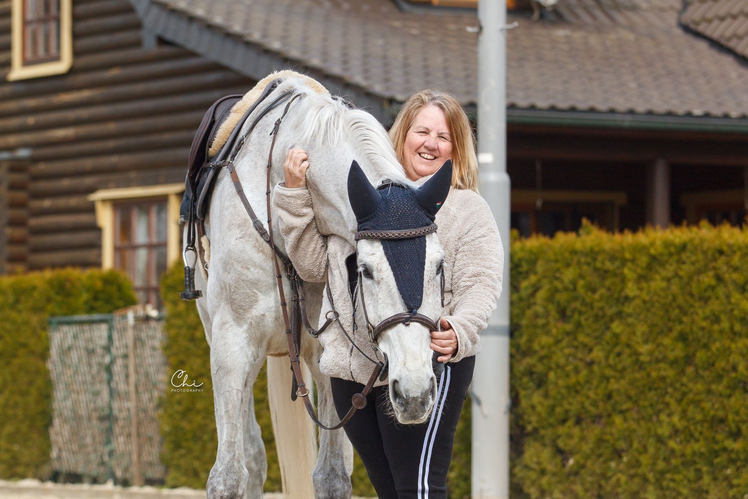 Happy woman standing beside grey horse outdoors near wooden building on sunny day.