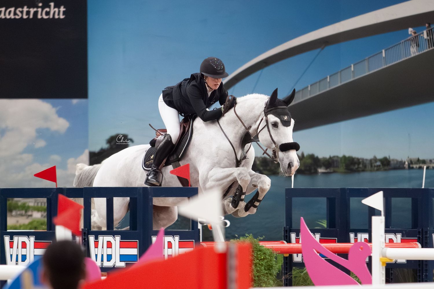 An equestrian rider jumps a grey horse over colorful obstacles at an outdoor show jumping competition.