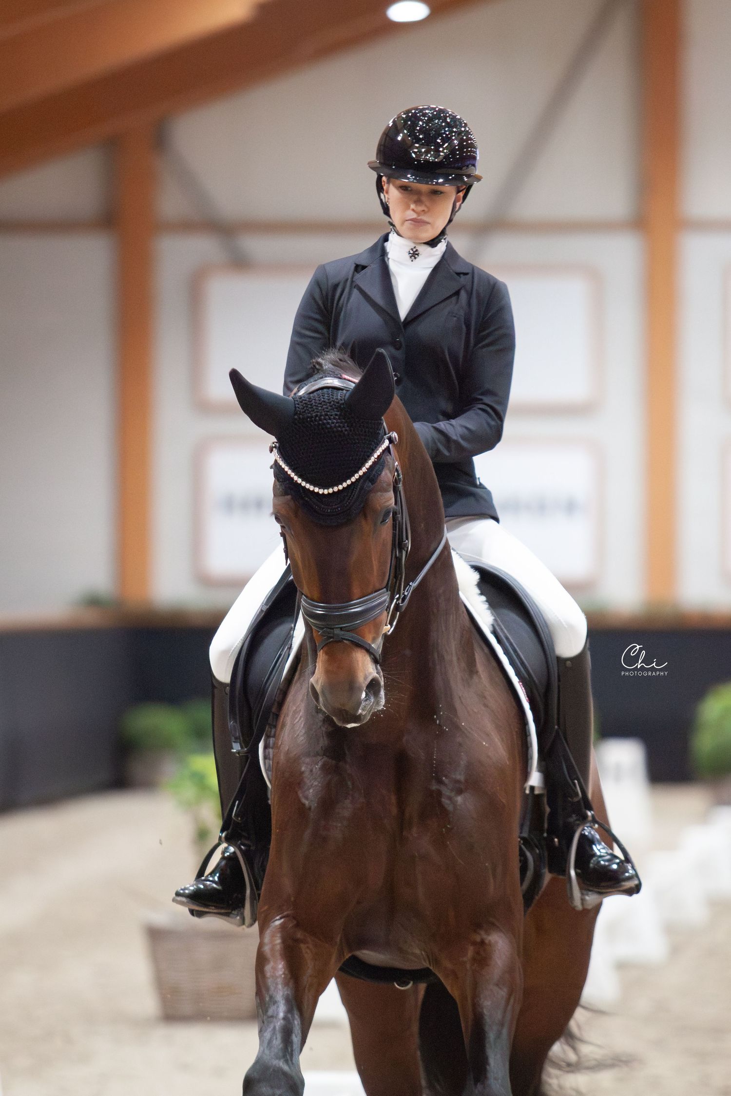 A focused young equestrian rider sits atop a bay horse wearing a sparkled helmet in an indoor arena.