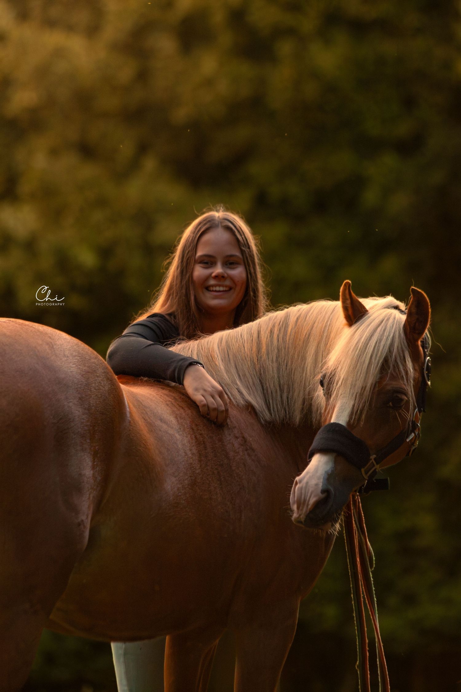 Smiling girl leaning on chestnut horse with blonde mane at golden sunset.