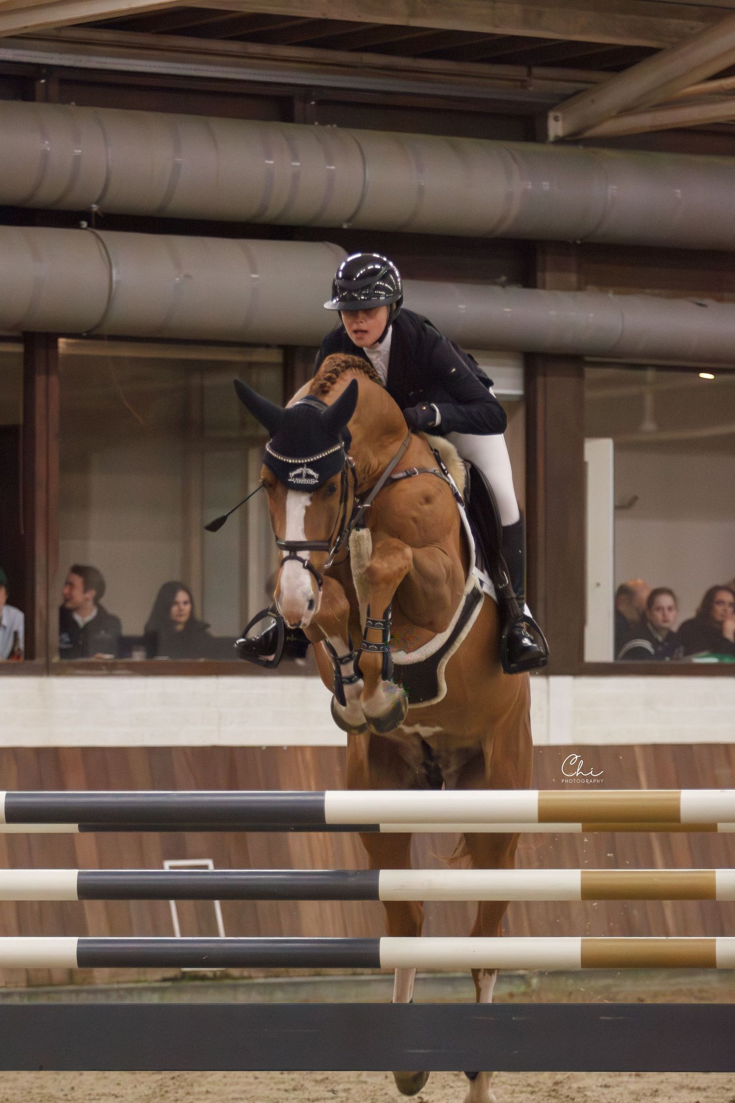 Show jumper on bay horse clearing colorful fence in indoor equestrian arena.