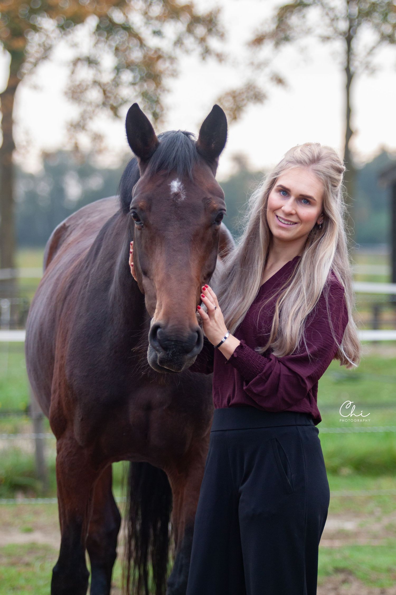 Smiling blonde woman in burgundy top feeding a dark bay horse in an autumn paddock setting.