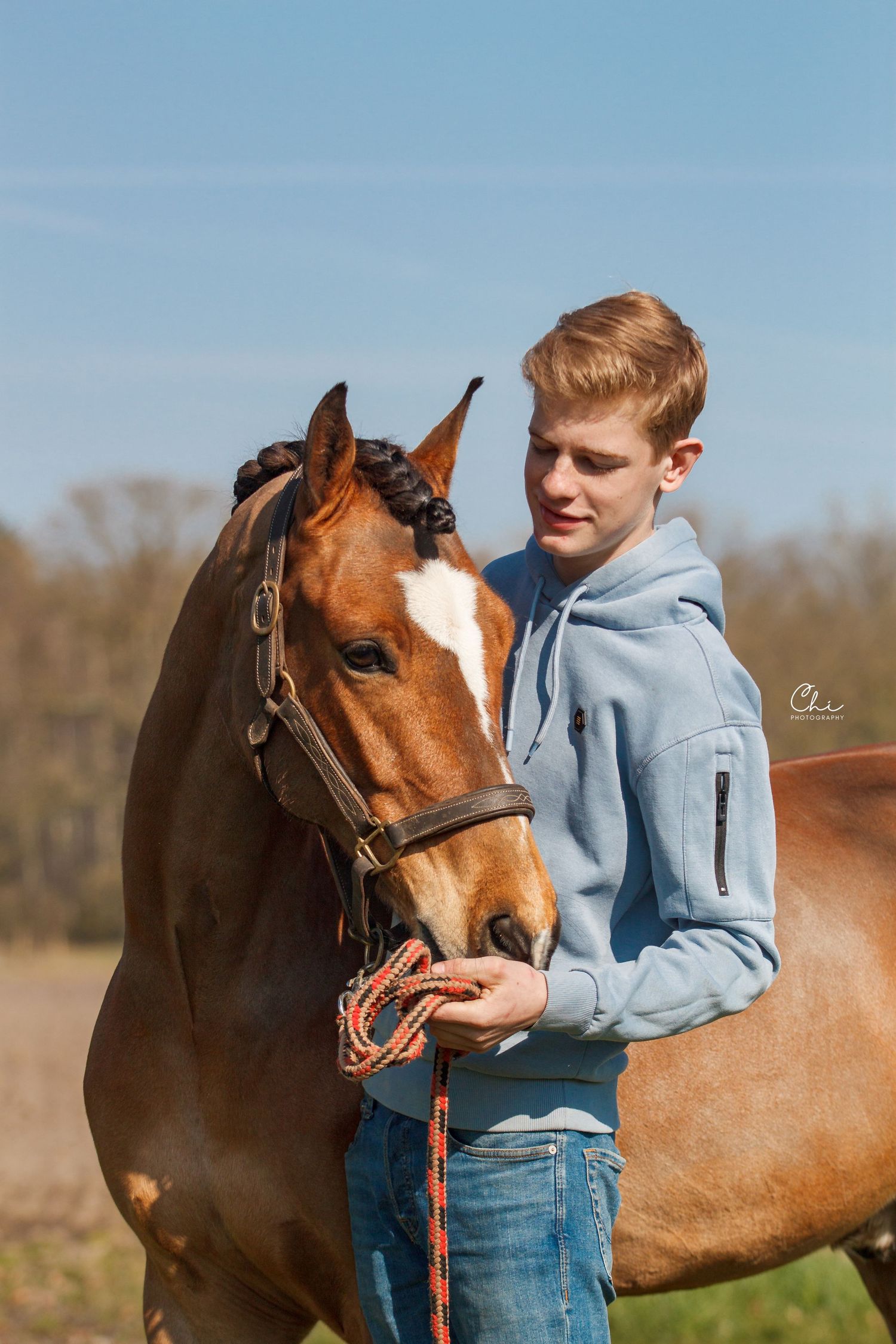 Young blond man in a grey hoodie gently holding a bay horse with a braided mane outdoors.