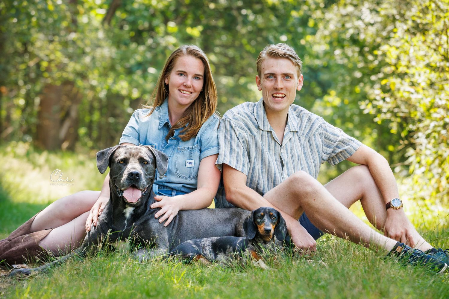 Young couple sitting on grass with two dogs, a large black dog and a small dachshund, in a sunny park.