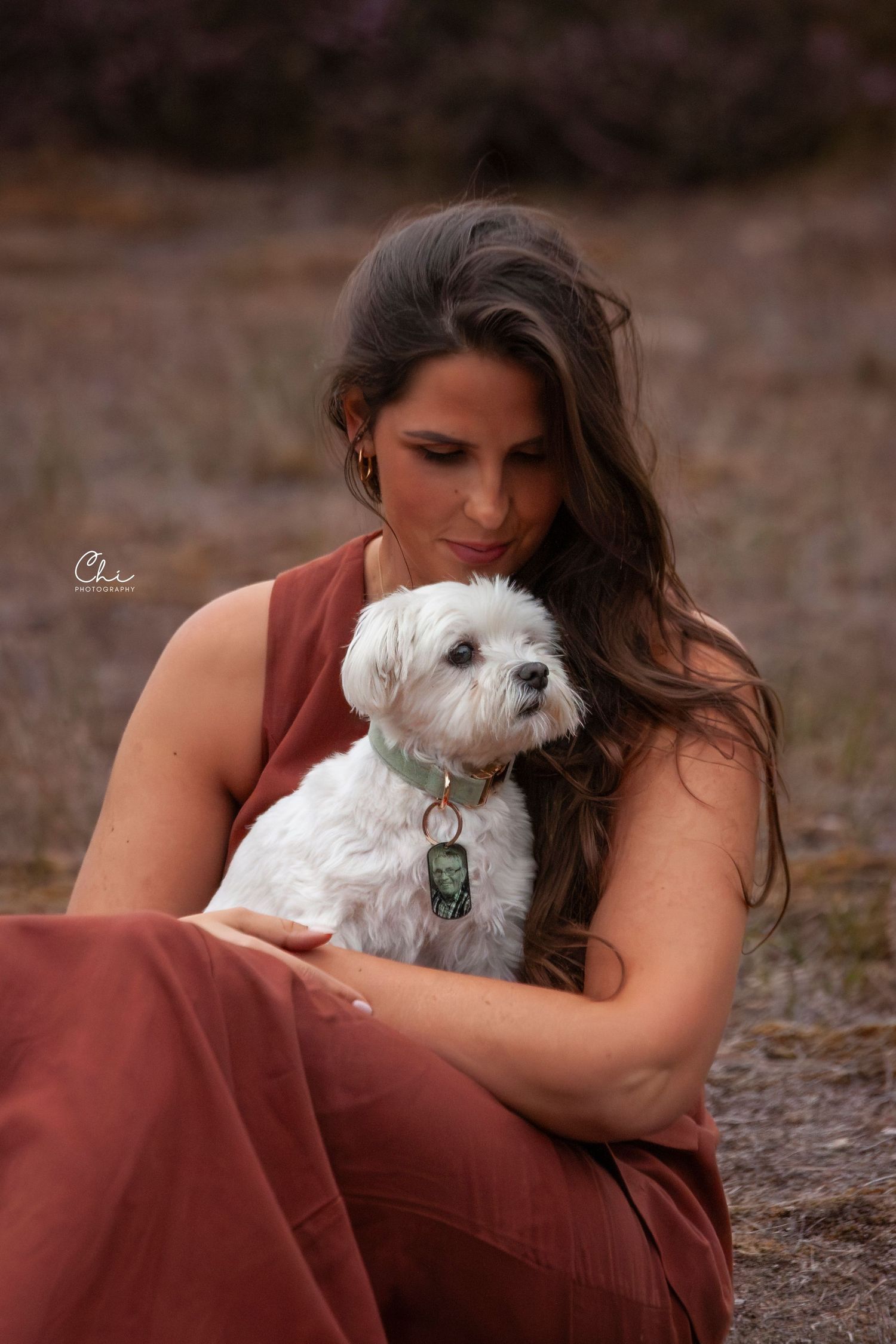 Young brunette woman in rust dress tenderly holding a small white Maltese dog.