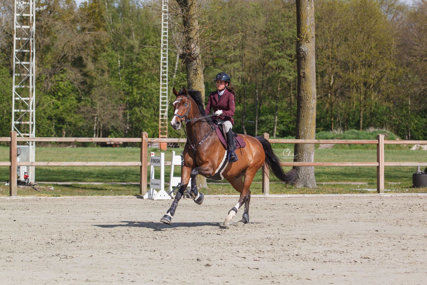 A rider in a maroon jacket and helmet canters a bay horse in an outdoor sand arena with wooden fencing.