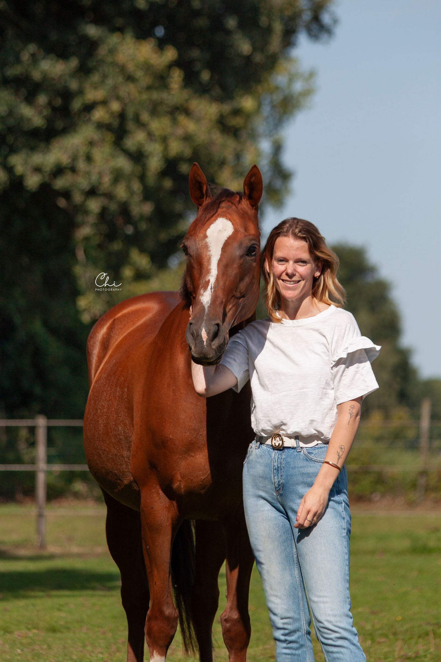 Smiling woman in white shirt standing beside chestnut horse in sunny field.