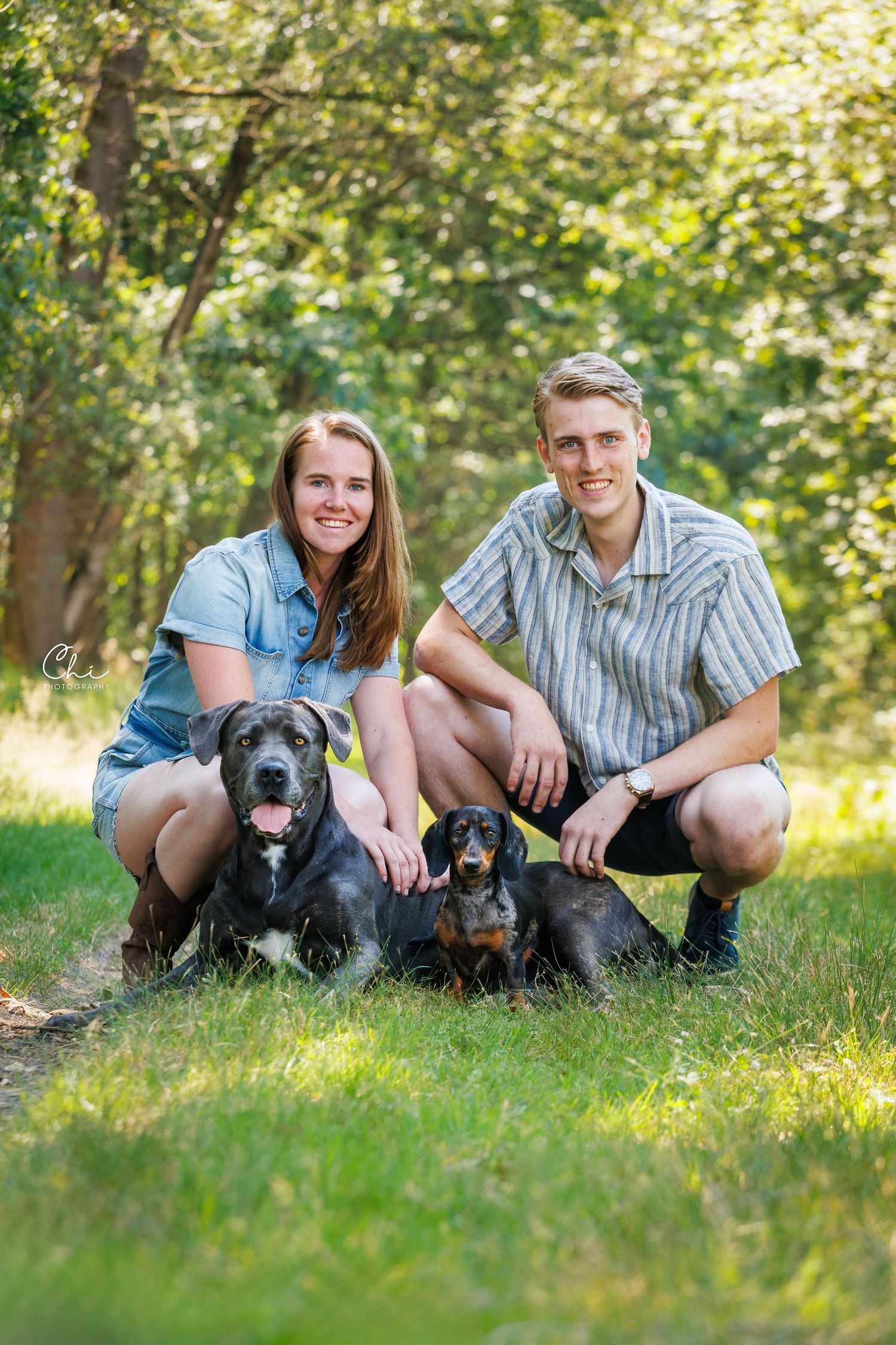 A smiling couple crouches on green grass with two black dogs, a large mixed breed and a dachshund, in a sunny park.