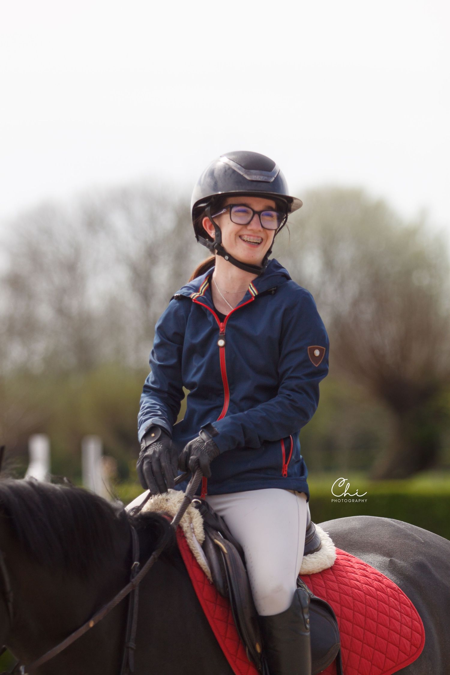 A smiling young girl wearing glasses and a riding helmet sits atop a black pony with a red saddle pad.