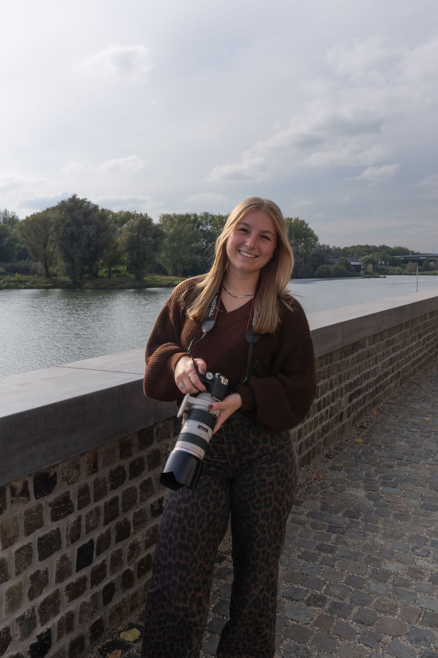 Smiling blonde woman holding professional camera on cobblestone bridge beside river.