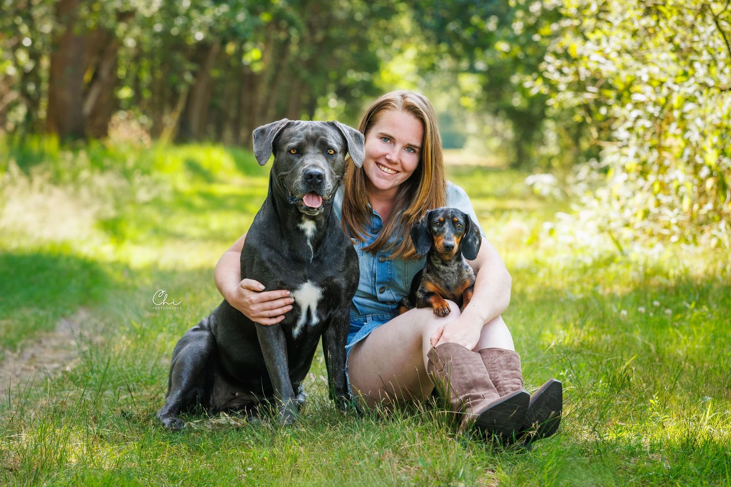 Woman smiling outdoors with a large black dog and a dachshund on a grassy forest path in natural light.