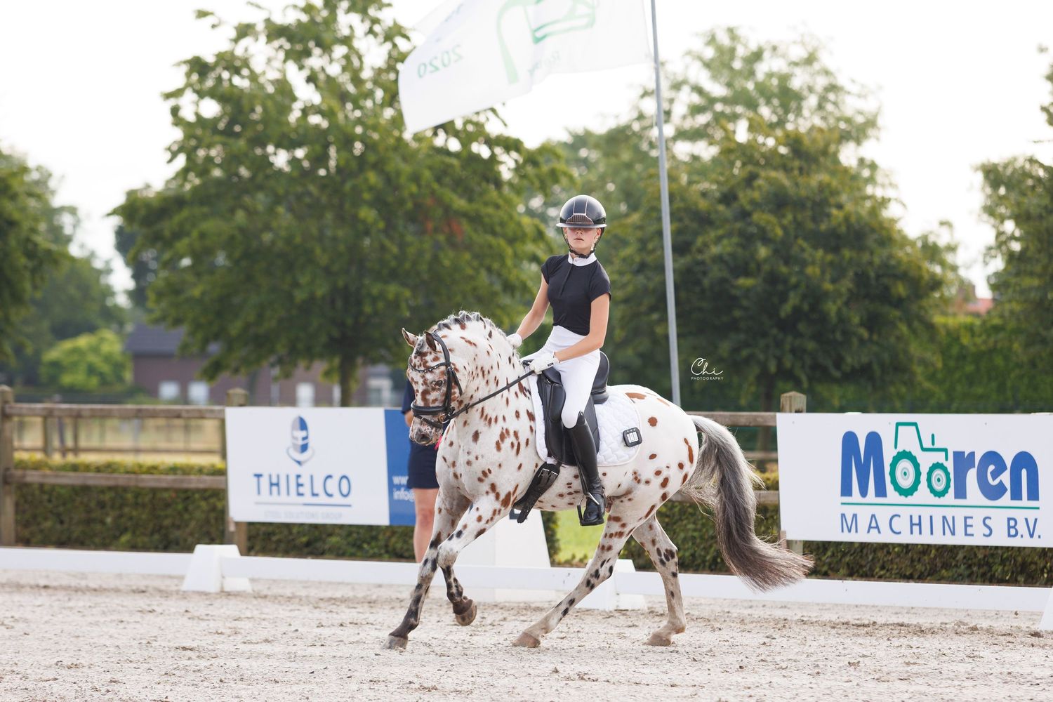 Young rider on spotted Appaloosa horse performing dressage at outdoor equestrian competition with sponsor banners.