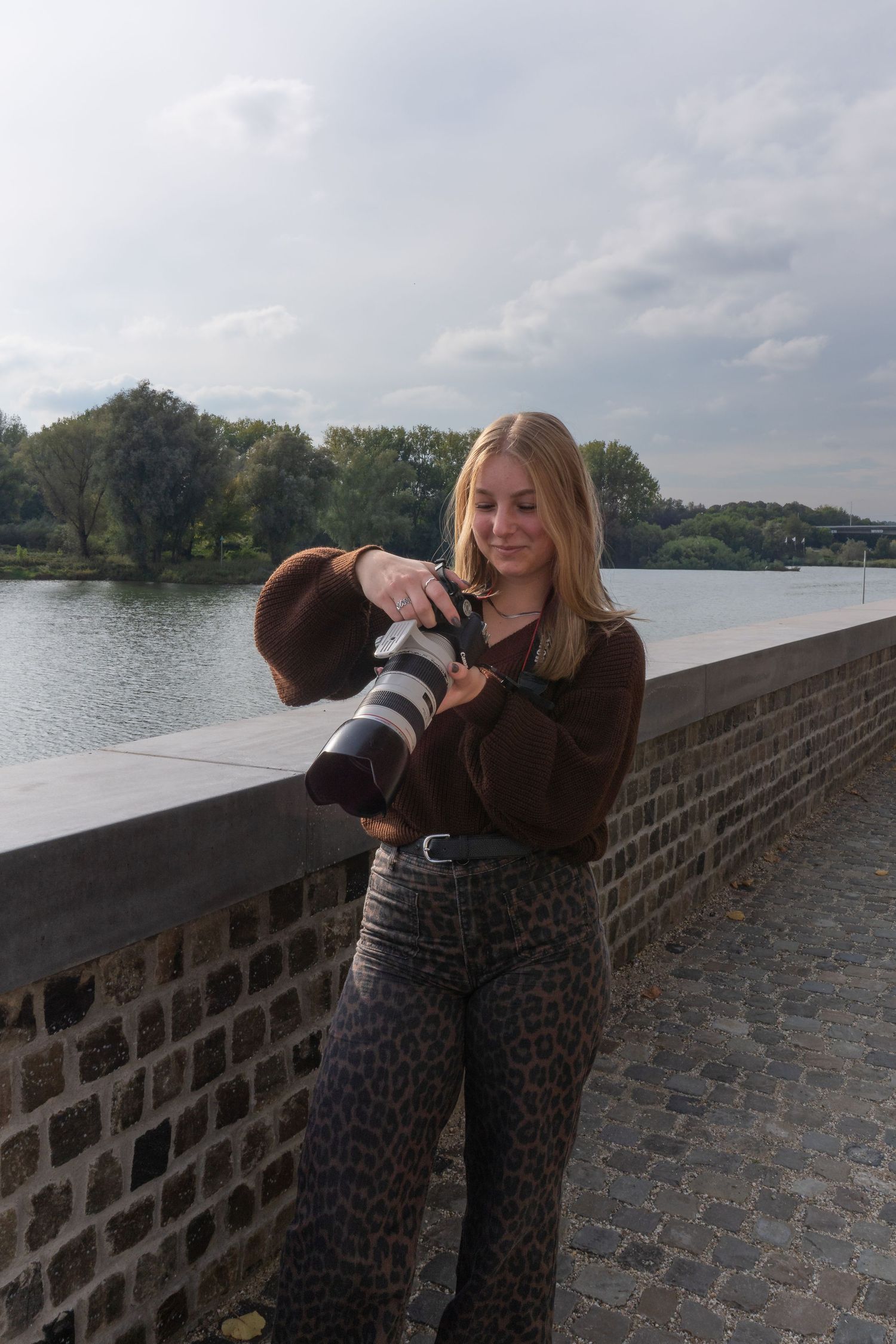 Blonde woman reviewing photos on professional camera while leaning on stone bridge.
