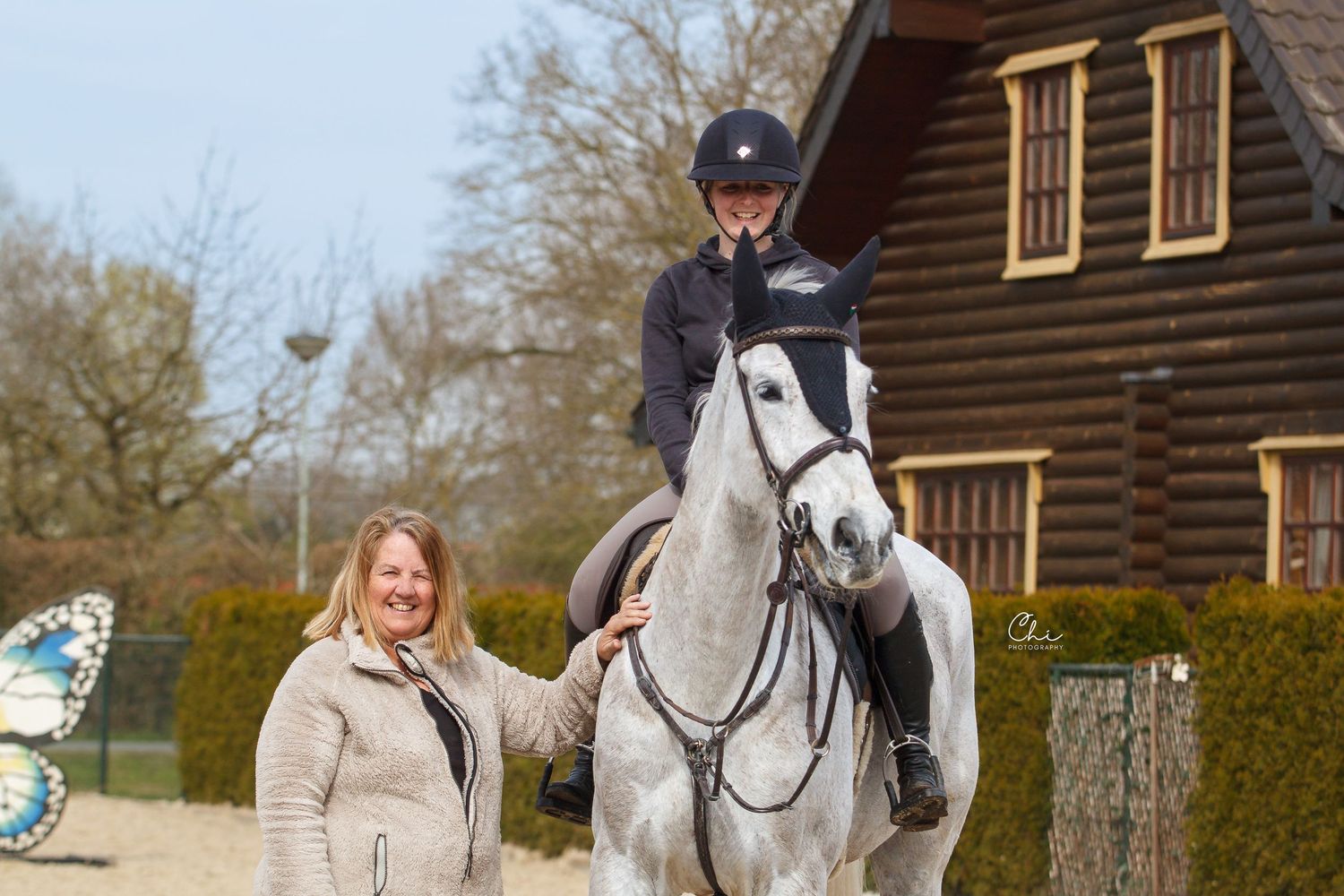 Woman standing beside rider on grey horse outside wooden equestrian building.