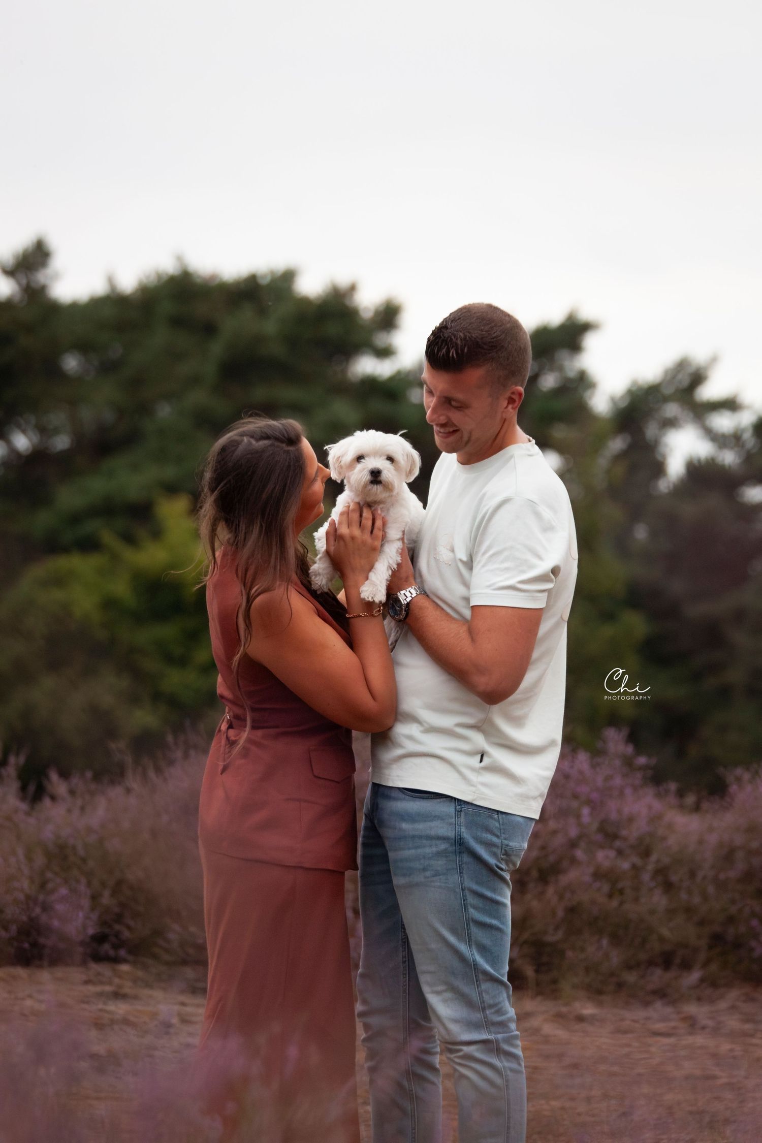 Smiling couple holding small white fluffy dog in purple heather landscape at dusk.