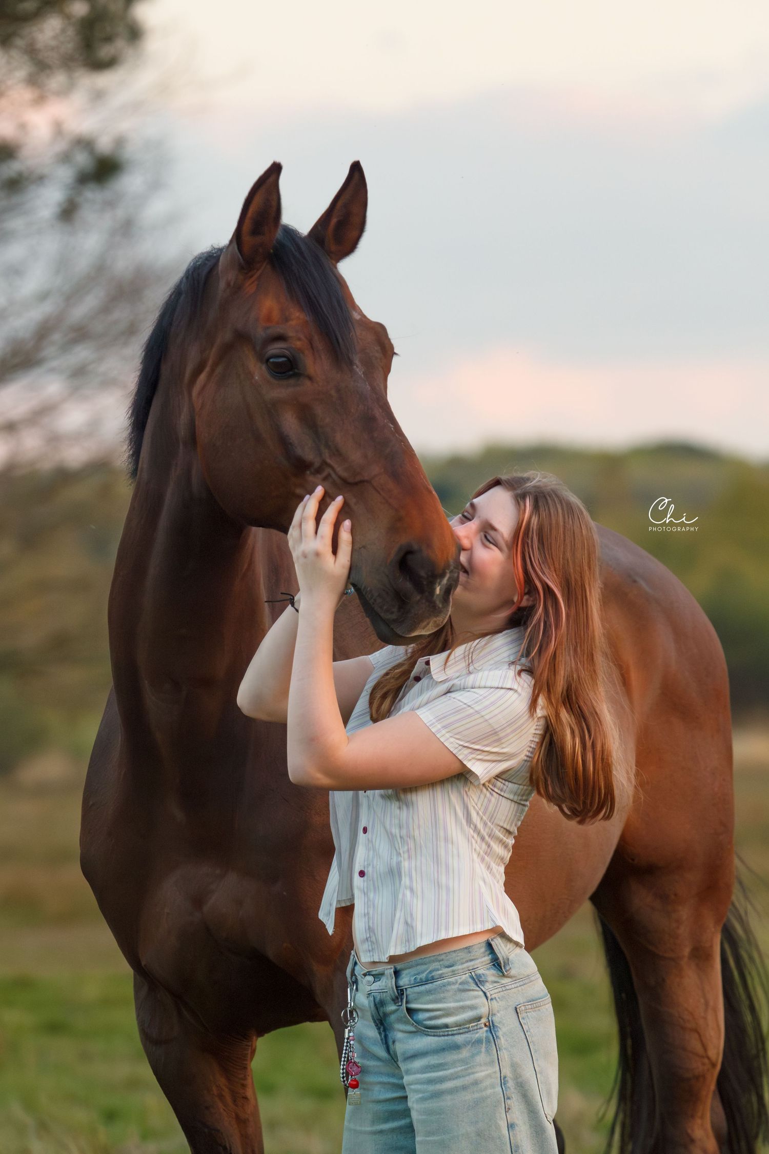 Young woman with long red hair affectionately kissing a bay horse's nose in a field at dusk.