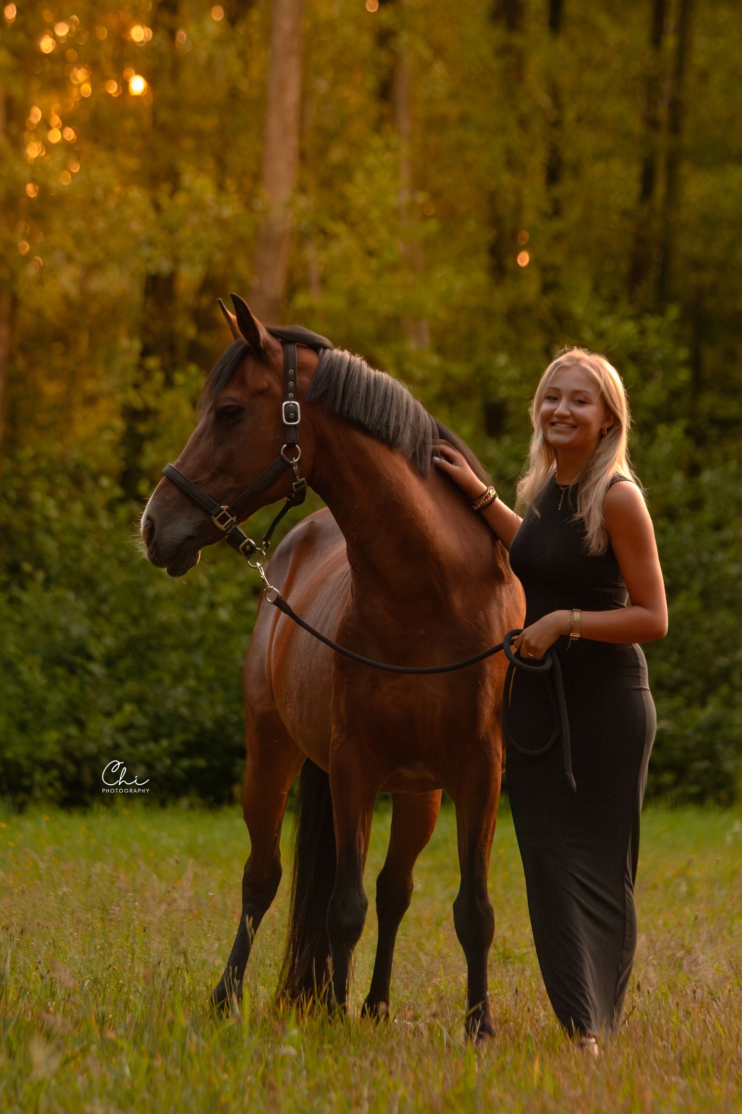 Smiling blonde woman in black dress standing with a bay horse in a golden-lit meadow with trees.