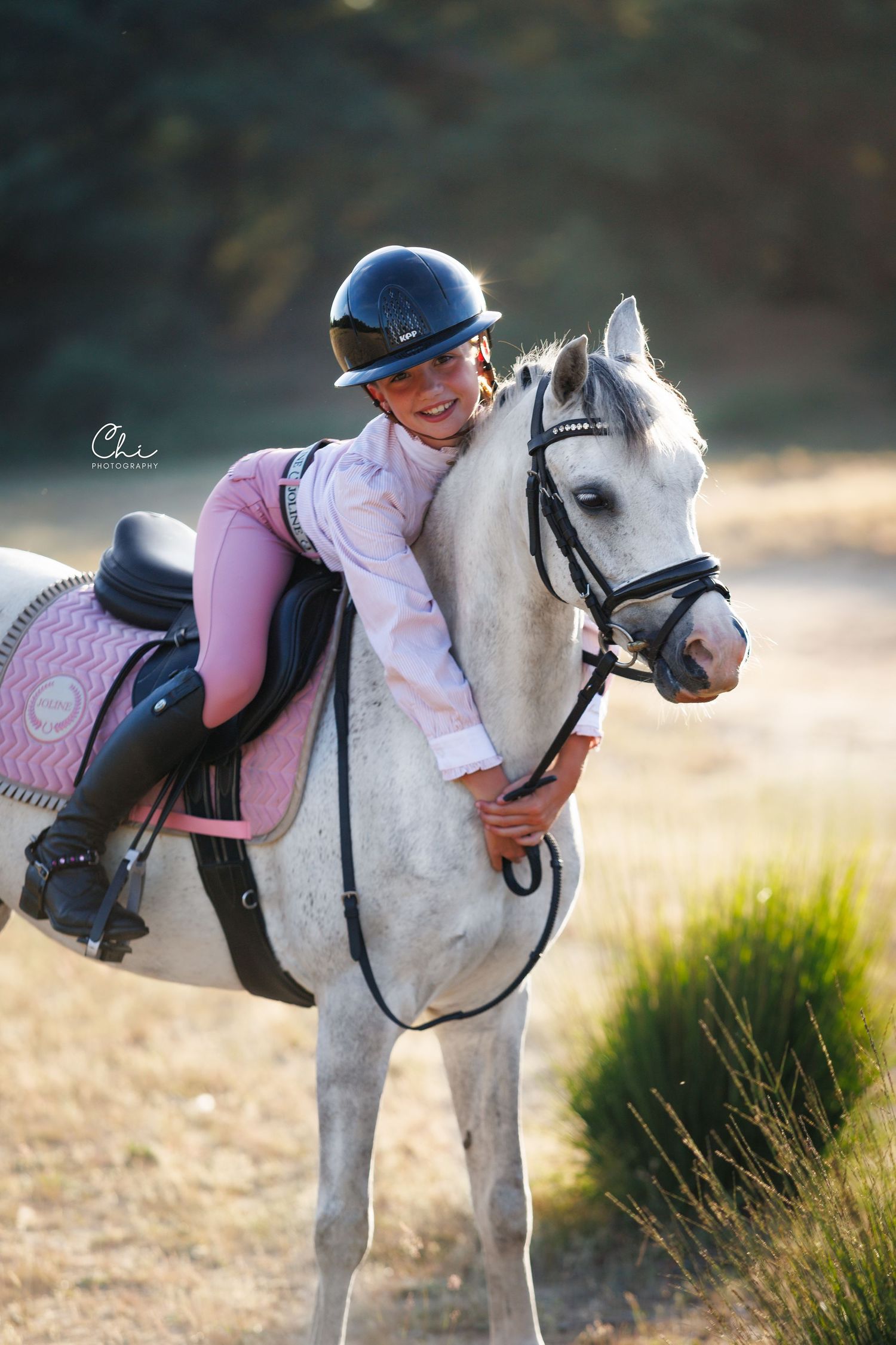 Smiling young girl in pink outfit and helmet hugging a white pony with pink saddle outdoors.