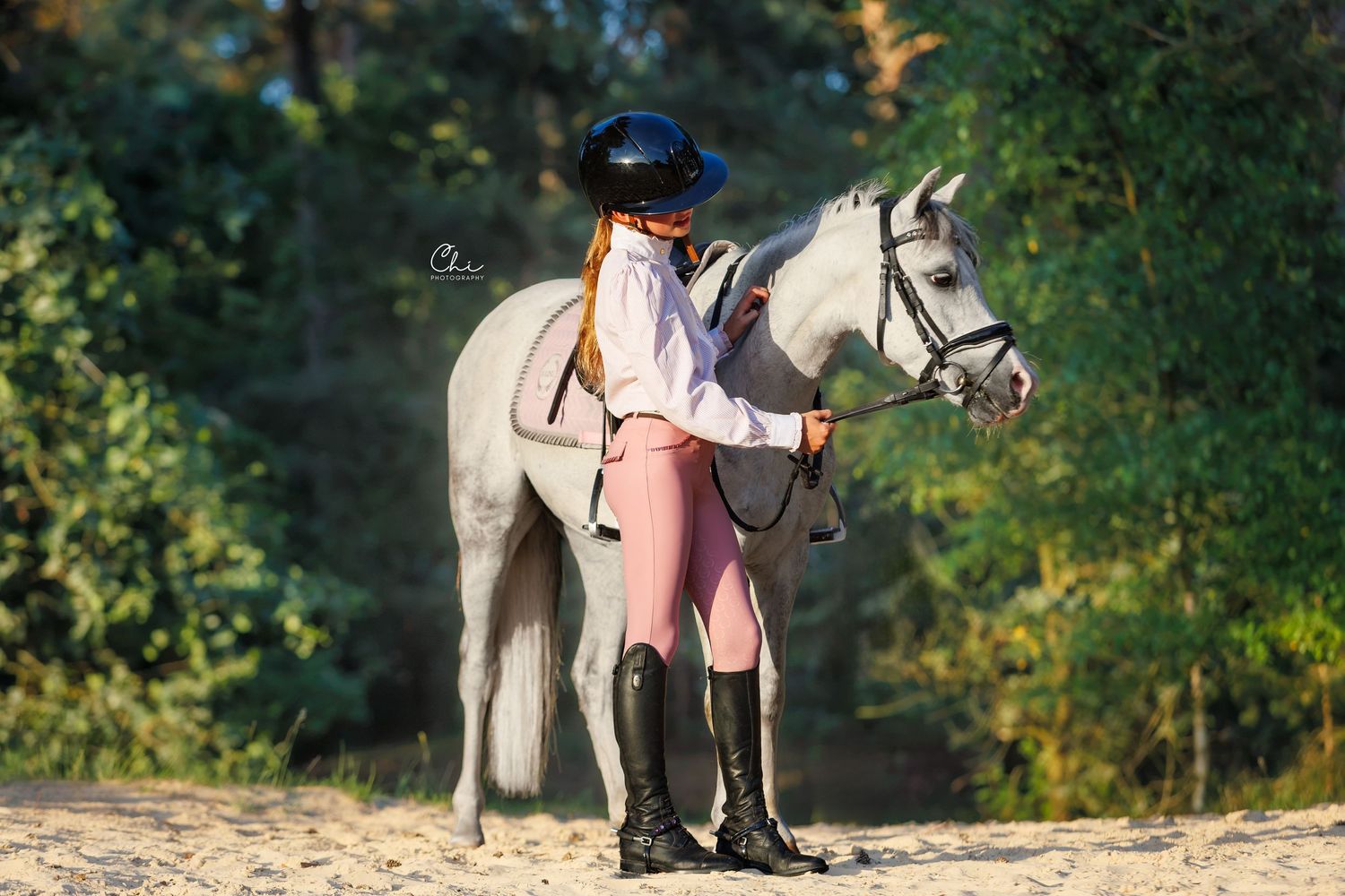 Young equestrian in pink breeches and black boots stands beside a white horse on sandy ground with green trees behind.
