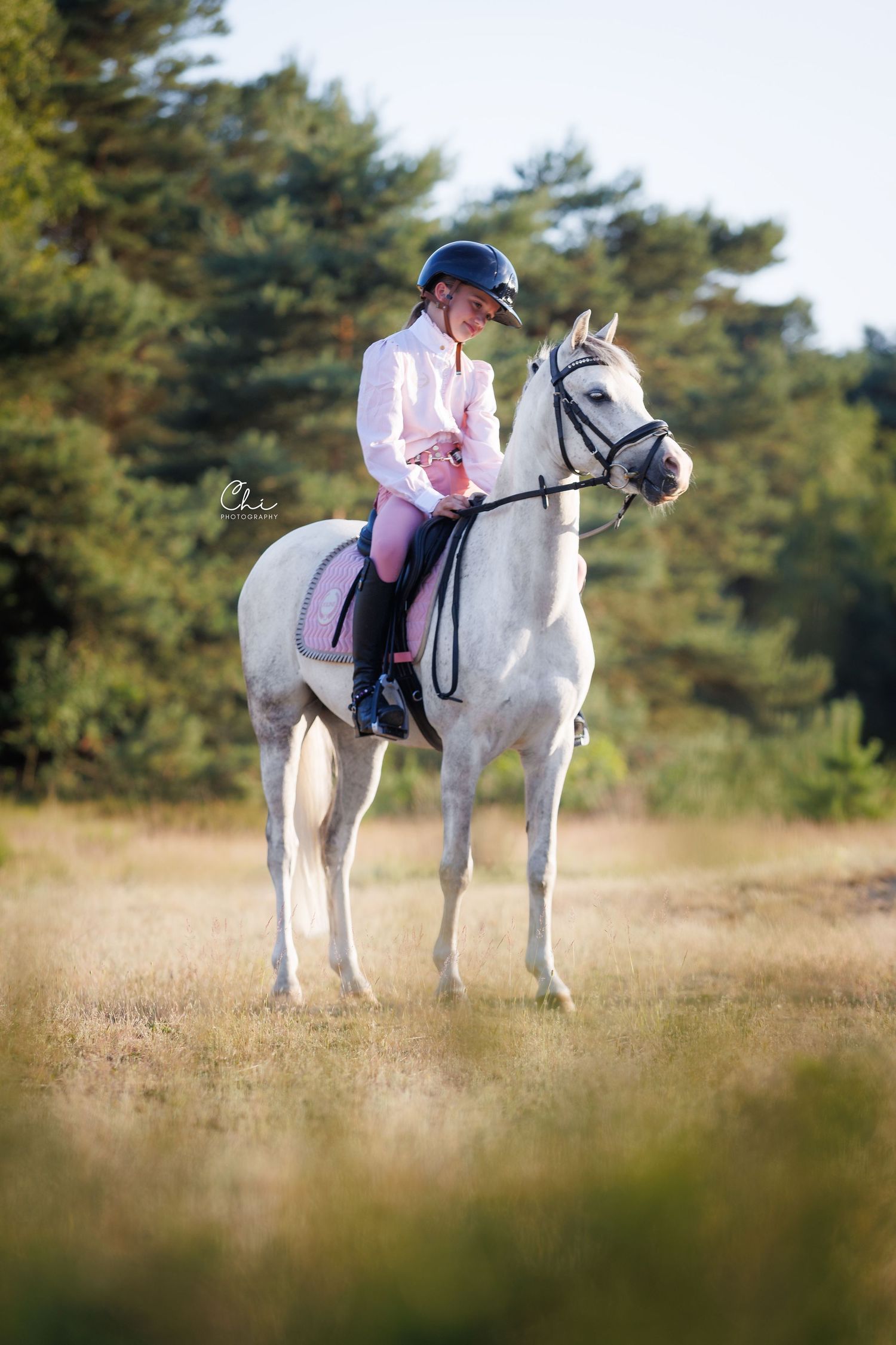 Young equestrian in pink and white riding a white horse in an open field with pine trees in the background.