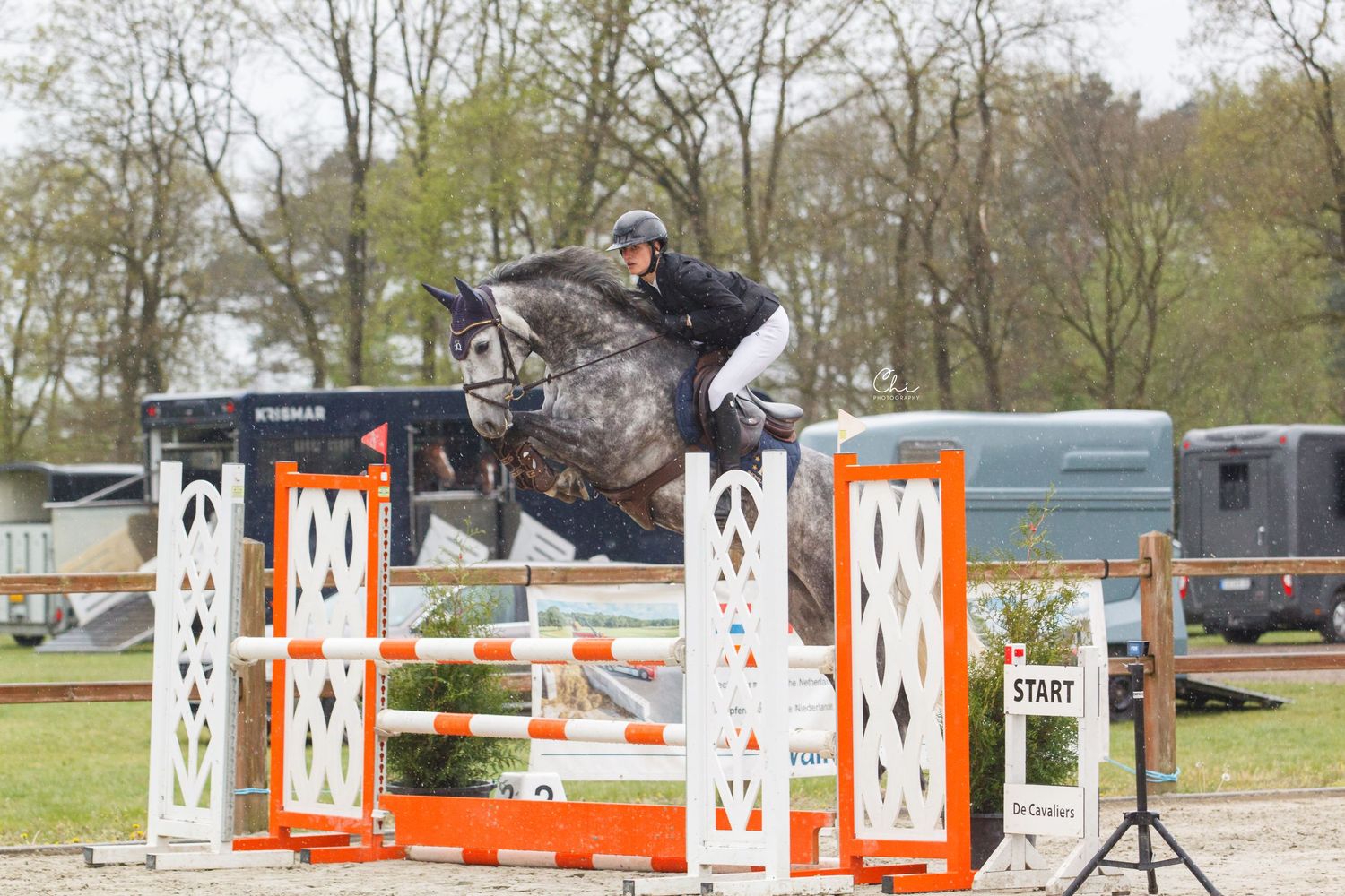 Equestrian rider on gray horse jumping over orange and white fence at outdoor show jumping competition.